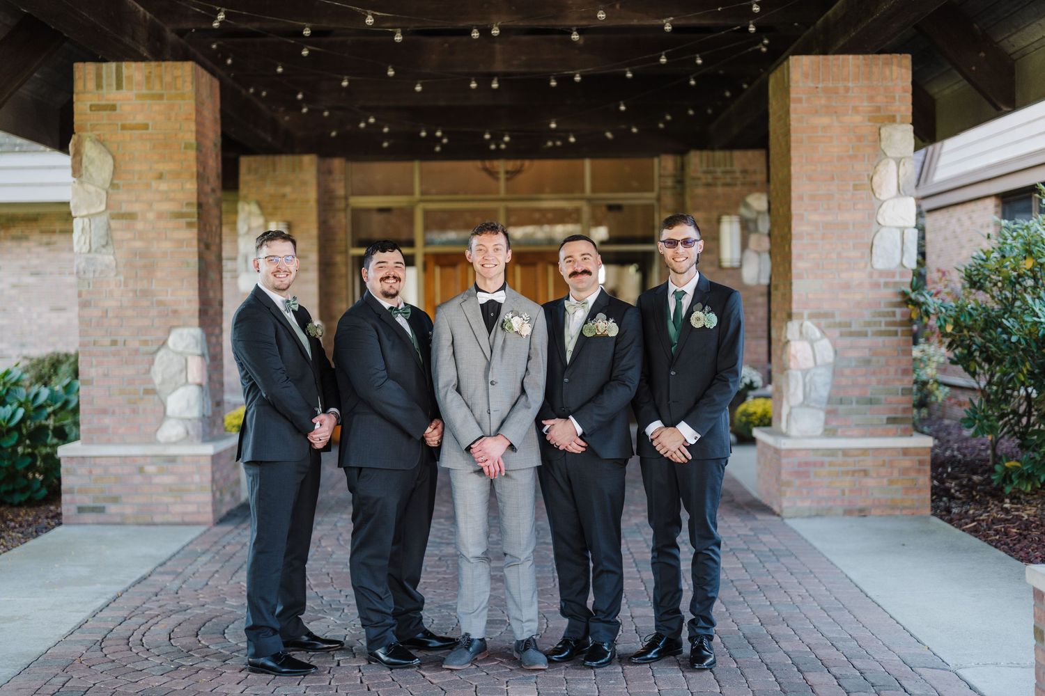 Groomsmen in dark suits line up on brick patio under string lights for formal wedding portrait.