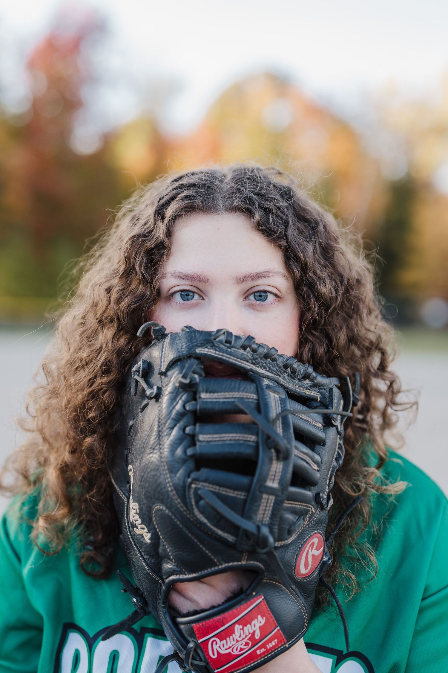 A person with curly hair wearing a green shirt playfully poses with a black baseball glove covering their face.