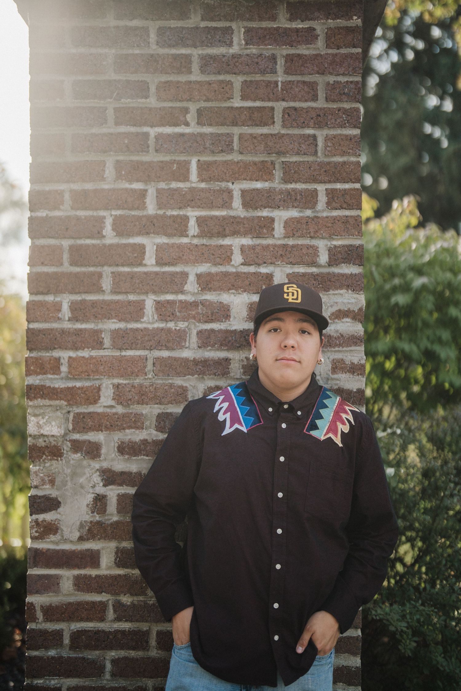 Person wearing a black button-up shirt with embroidered details poses against a brick wall outdoors.
