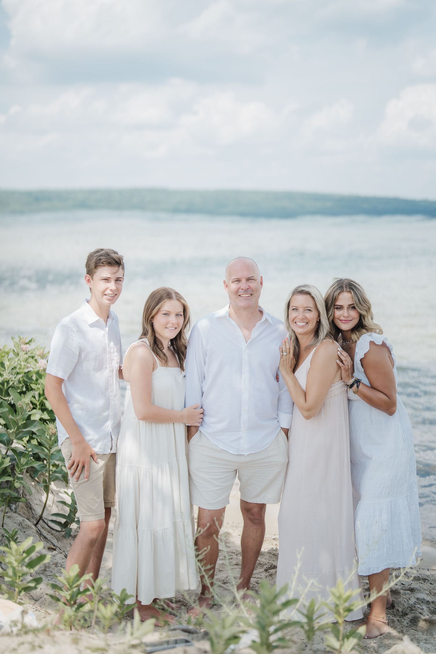 A family group wearing coordinated white outfits poses together on a scenic beach with ocean views in the background.