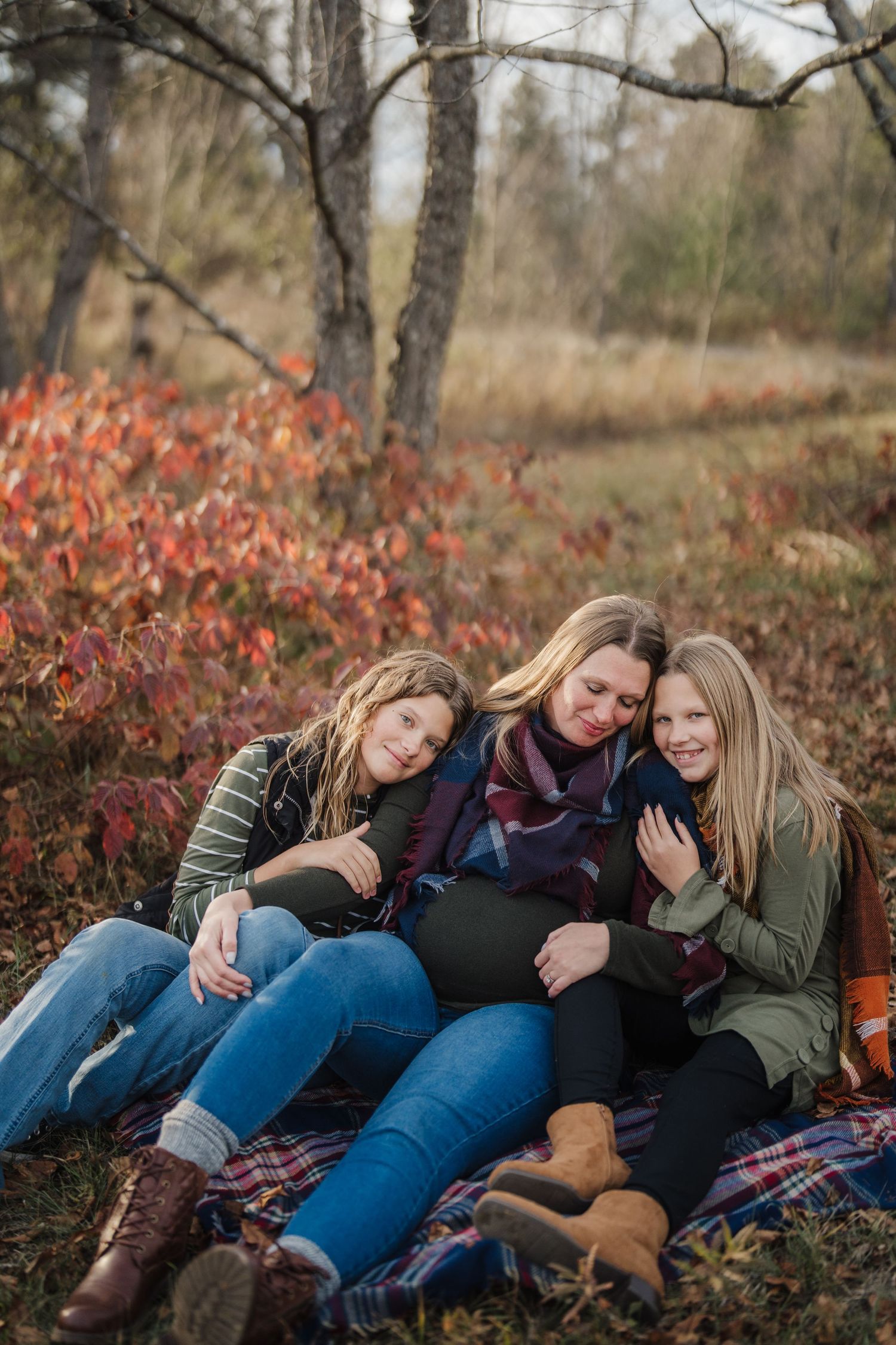 Three people relax together on a blanket outdoors during autumn with red foliage in the background.