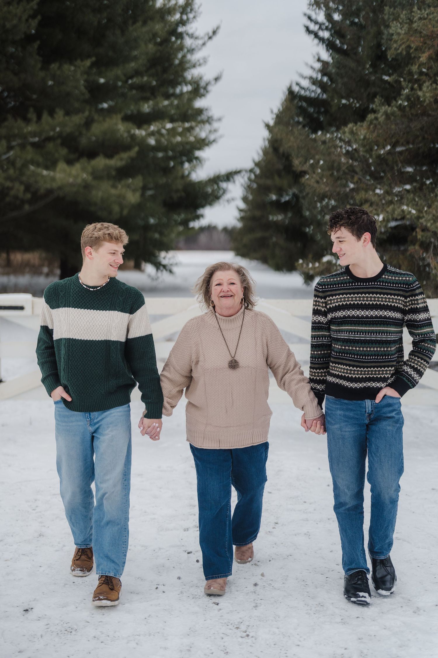 Three people walking together on a snowy path surrounded by evergreen trees wearing cozy winter sweaters.