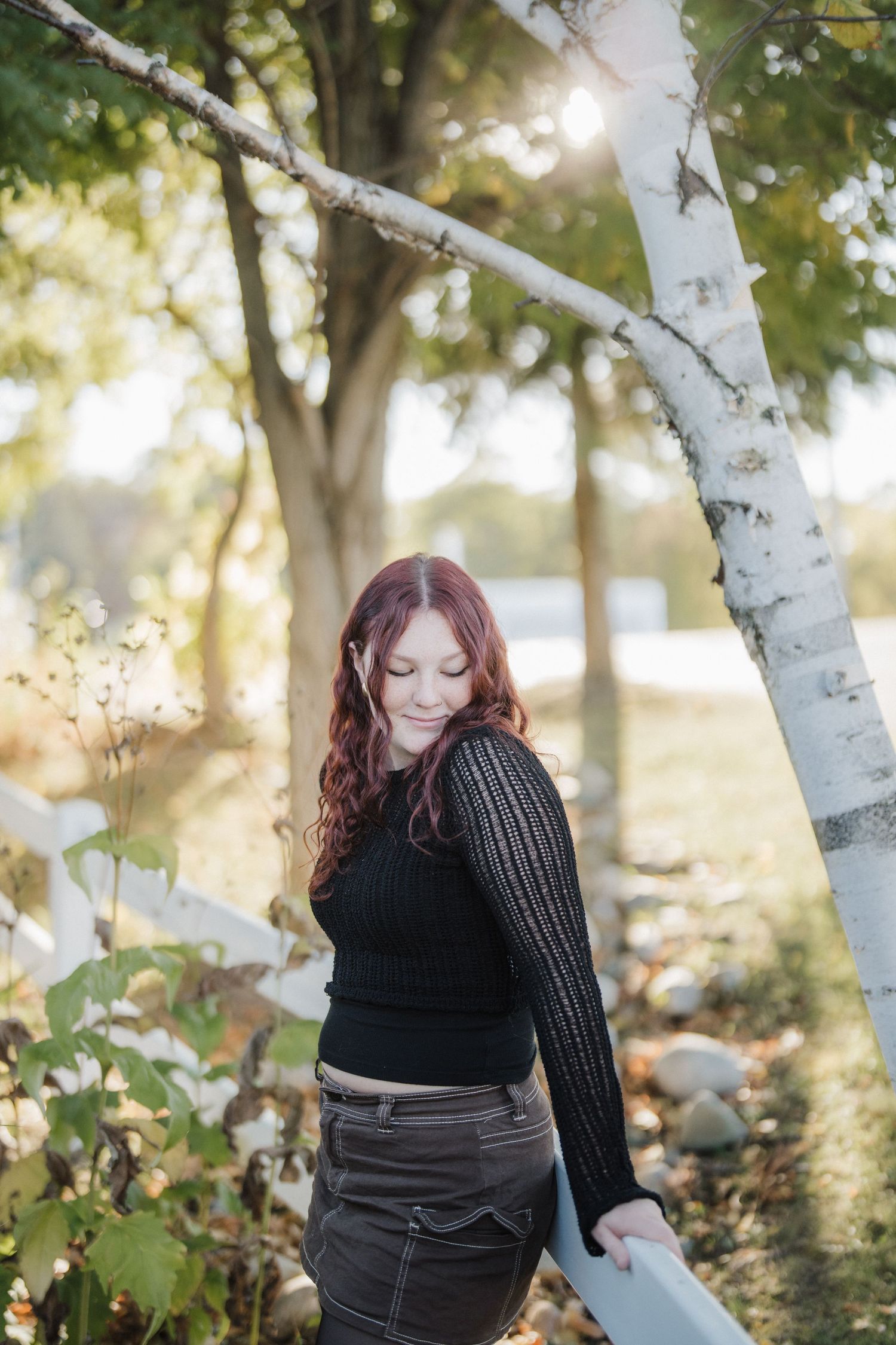 Person in dark sweater sits thoughtfully near white birch trees in a sunlit outdoor setting during autumn.