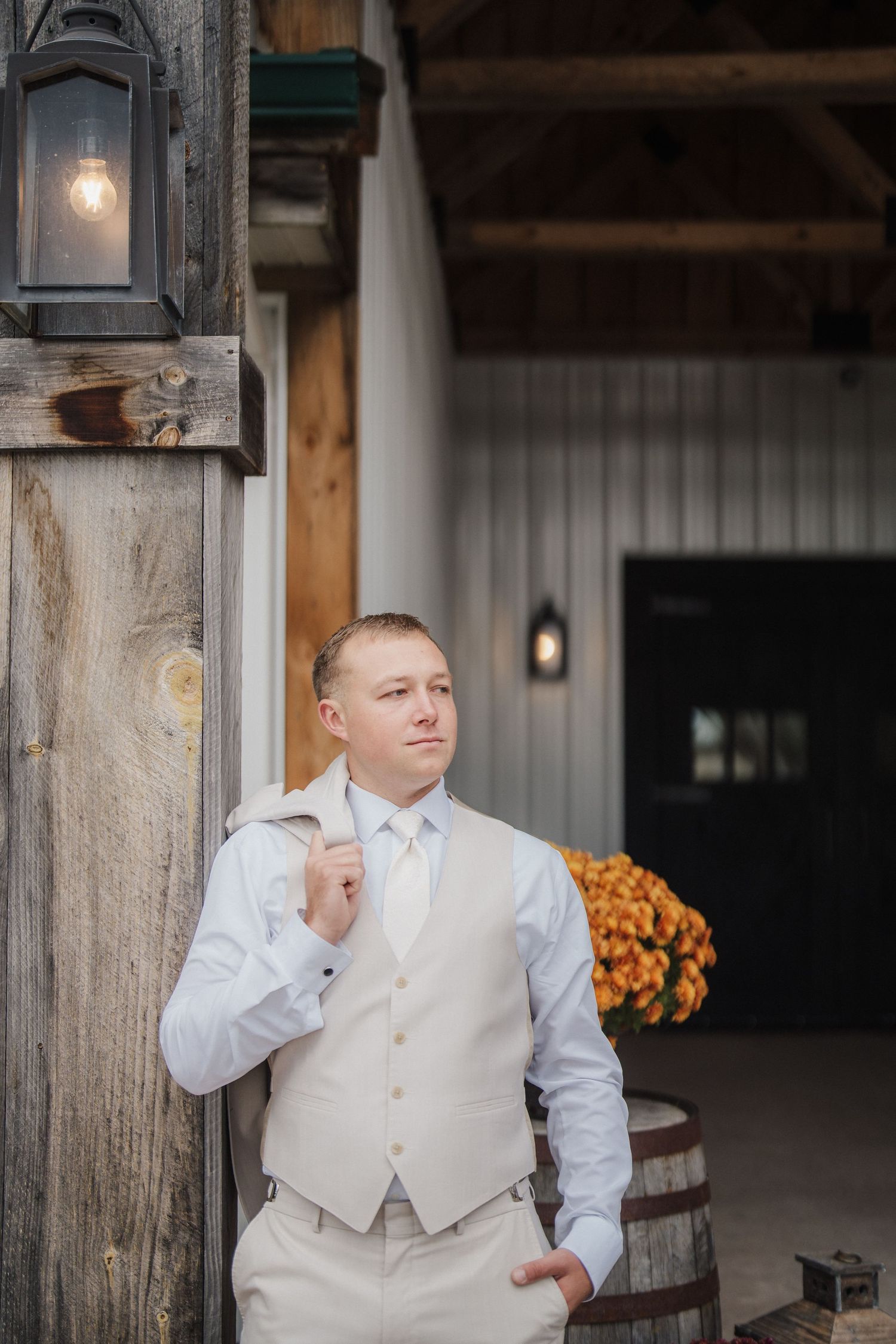 Groom in light-colored vest and white shirt stands thoughtfully in rustic venue setting.