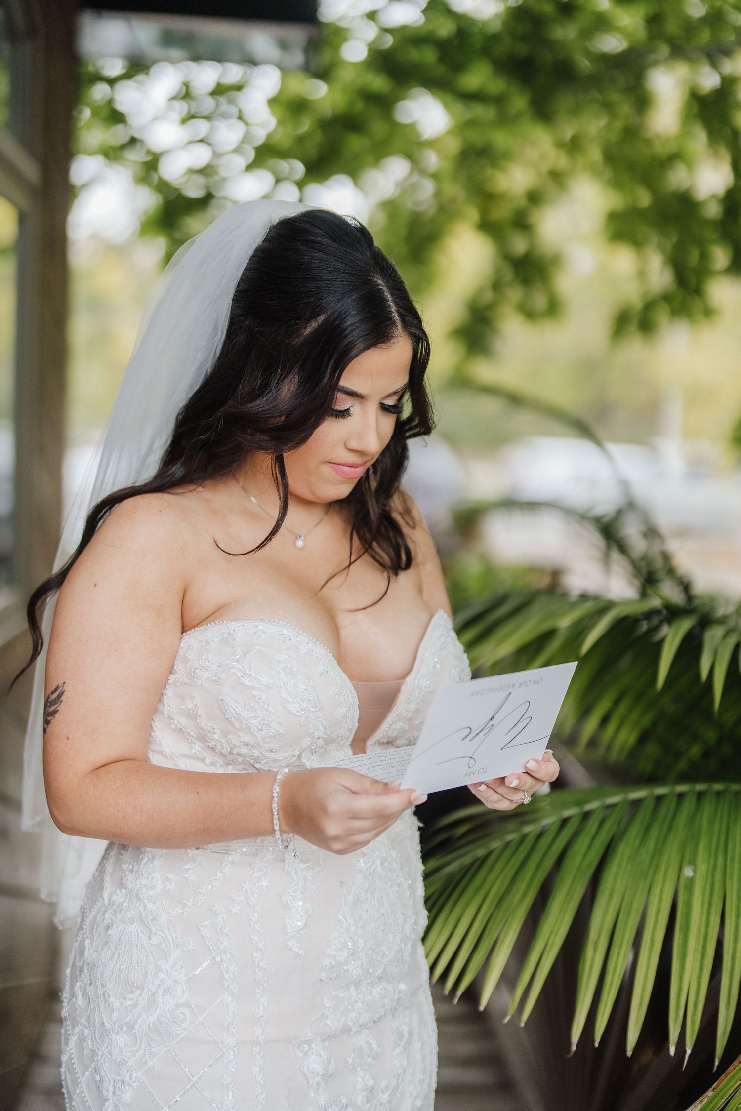 Bride in a strapless lace gown reading a letter near tropical palm leaves.