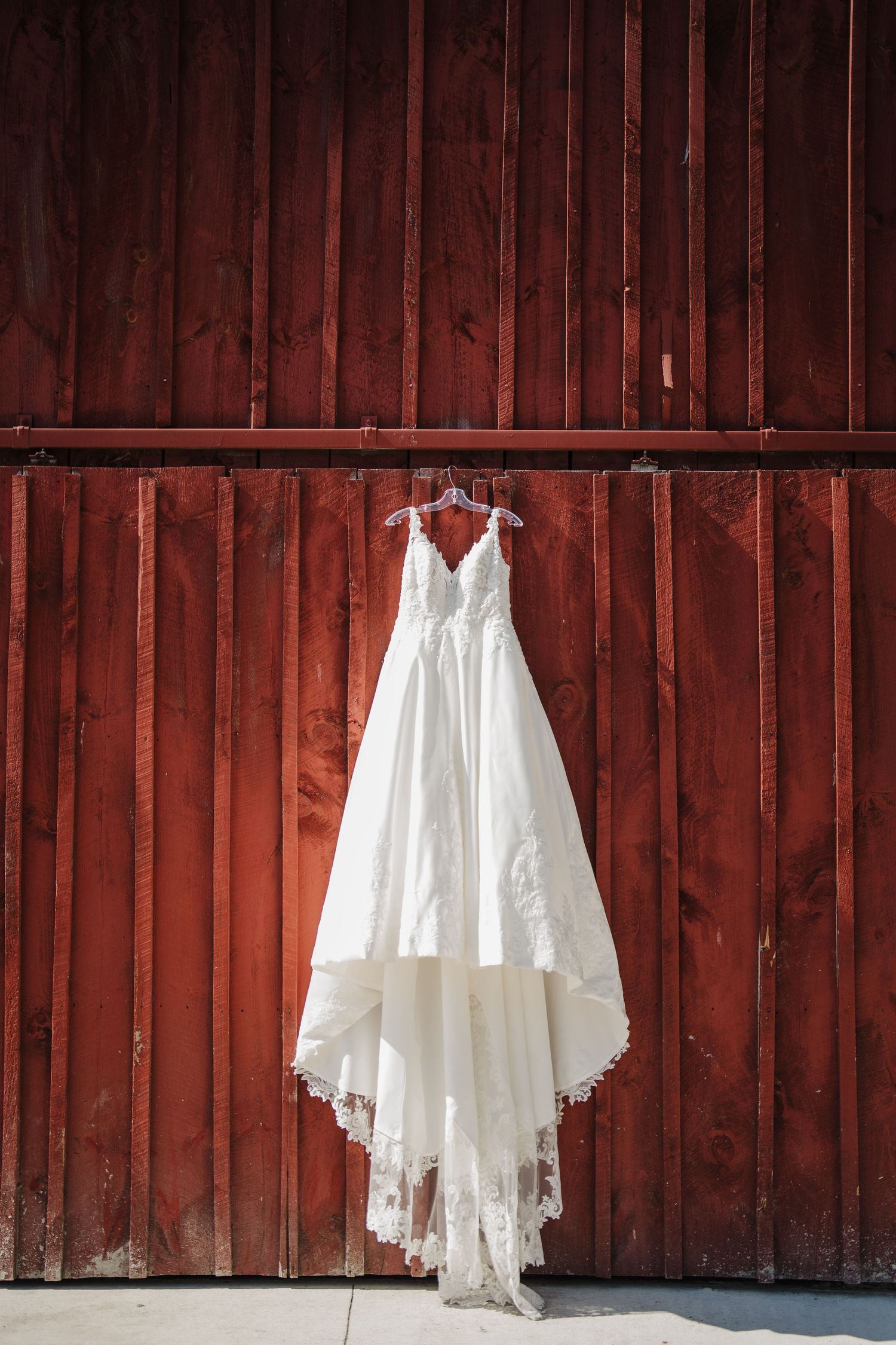 A white lace wedding dress with a long train hangs against a rustic red barn wall.