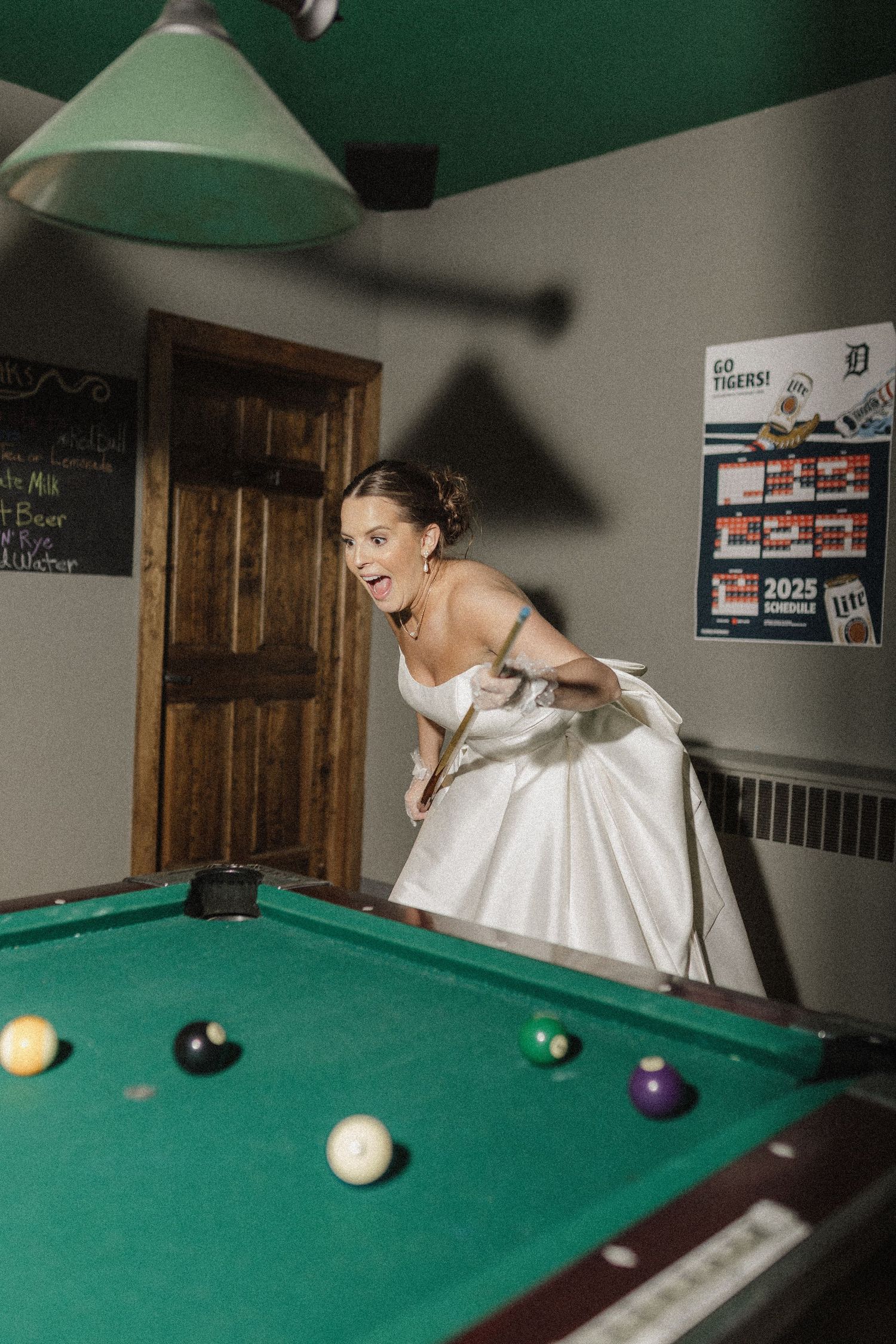 A bride in a white wedding dress plays pool in a game room, leaning over the table to take a shot.
