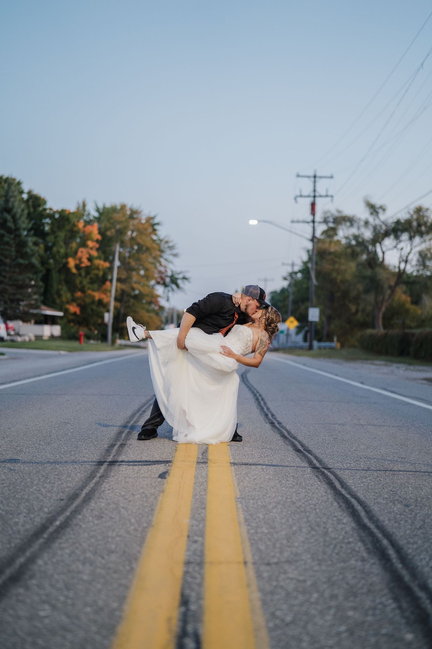 A couple poses for creative wedding photos lying on a rural road with double yellow lines at sunset.