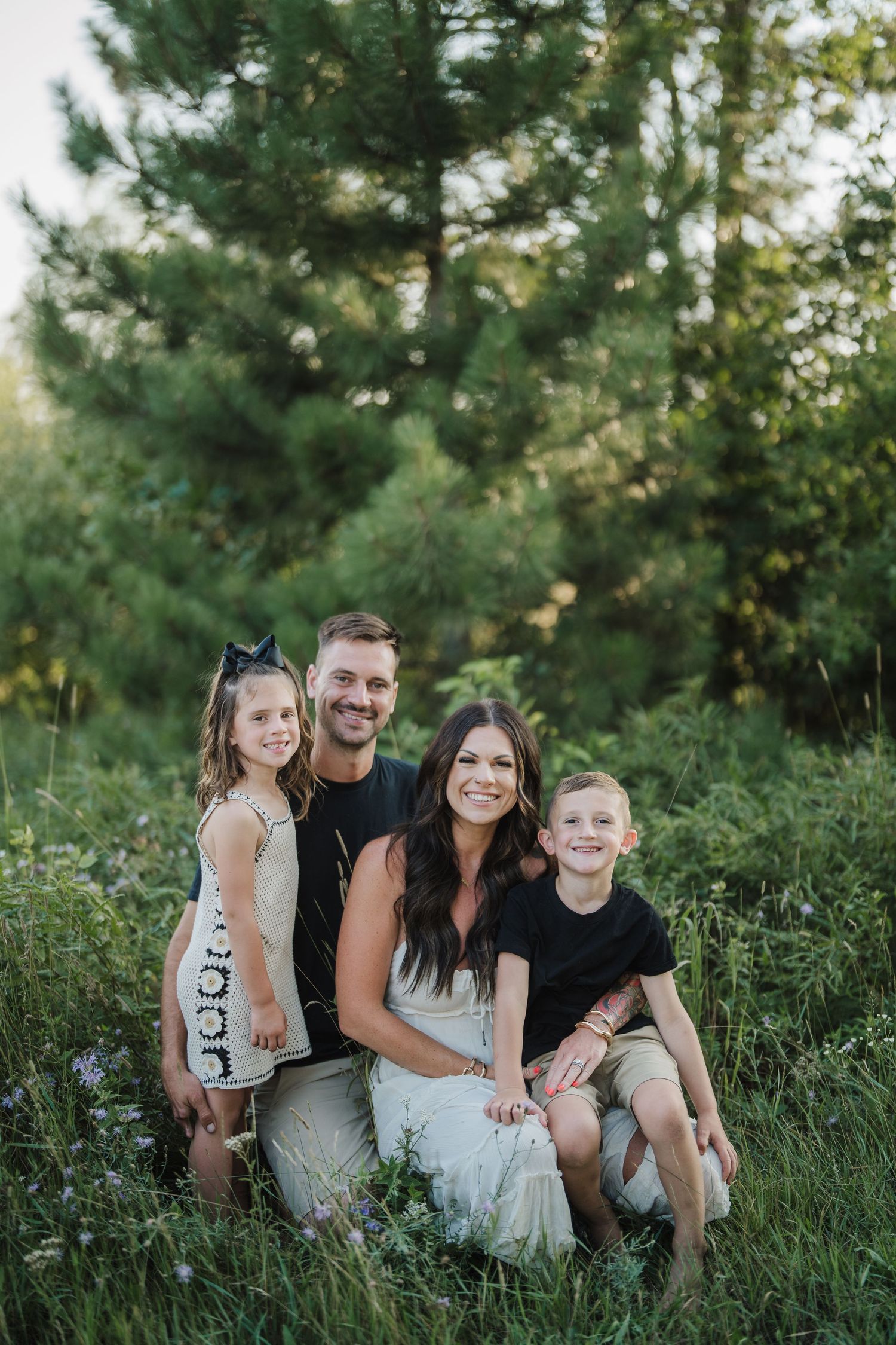 A family sits together on grassy ground surrounded by green trees during a casual outdoor photo session.