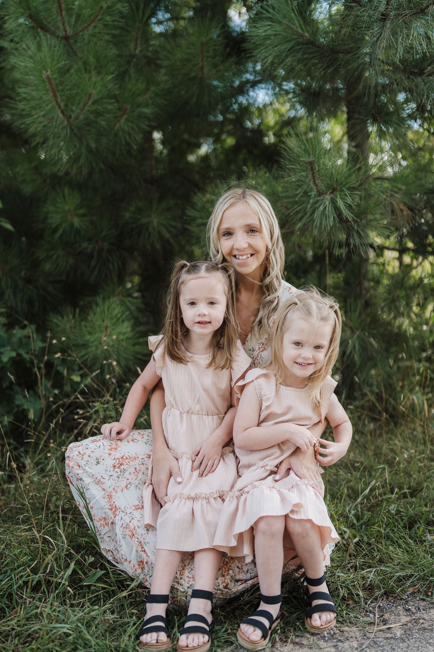 Three people in matching pink dresses sit together against a backdrop of green pine trees on a sunny day.