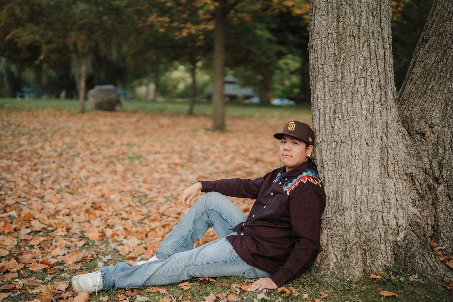 Person relaxing against a tree trunk in a park covered with fallen autumn leaves during golden hour.