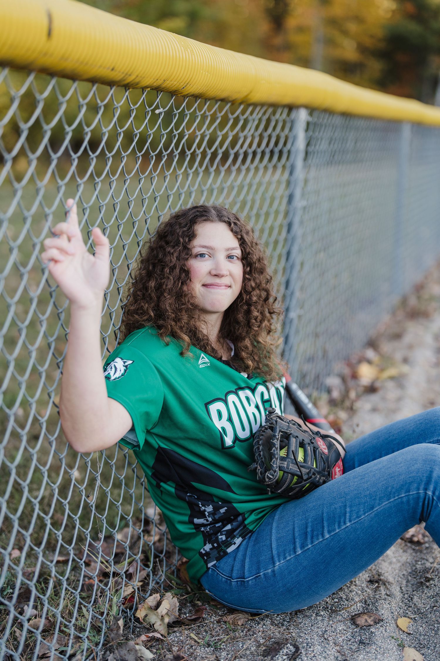 Person in green jersey poses casually by yellow railing and chain link fence during fall season.