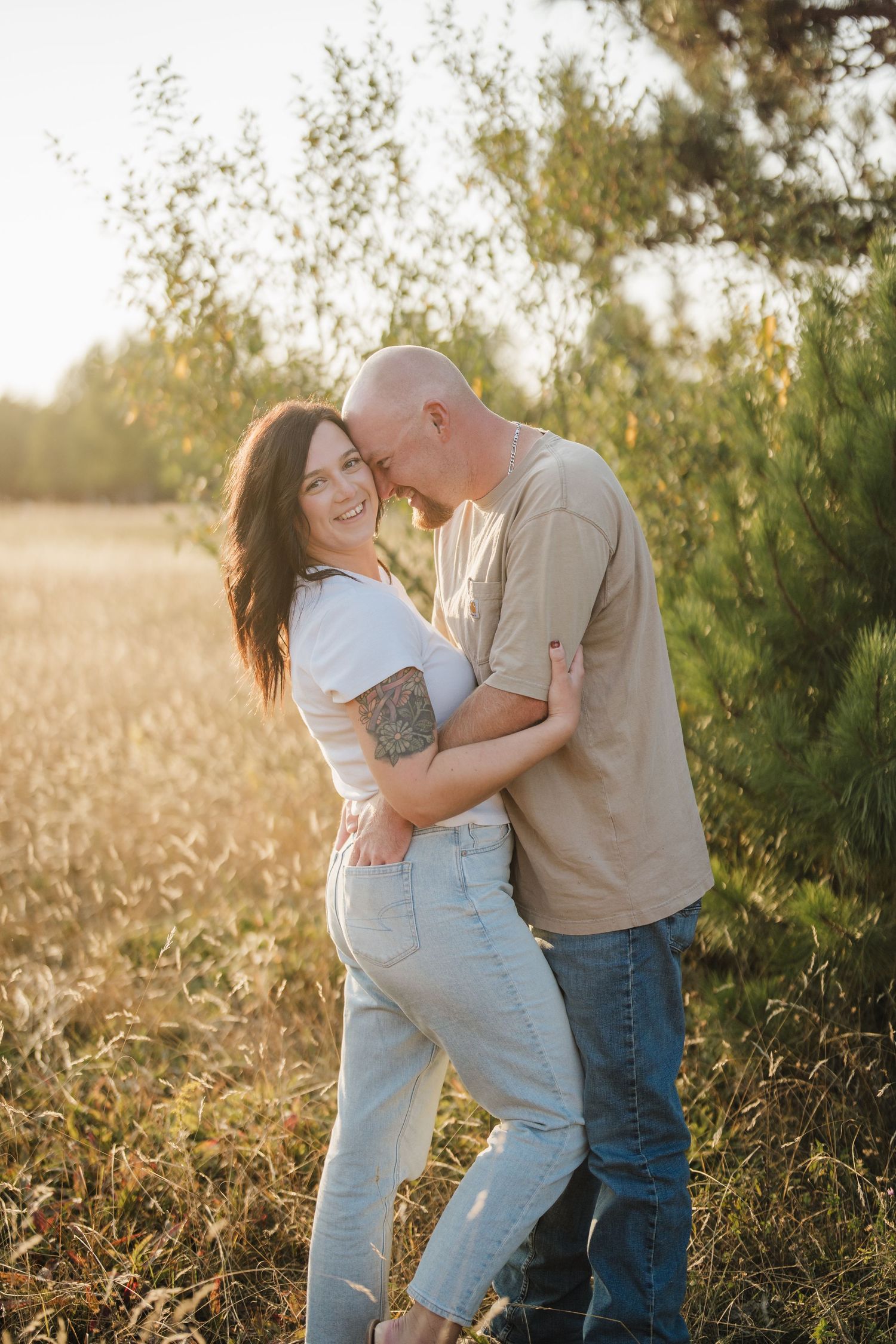 A romantic couple embraces and laughs together in a sunlit field at golden hour with trees in the background.