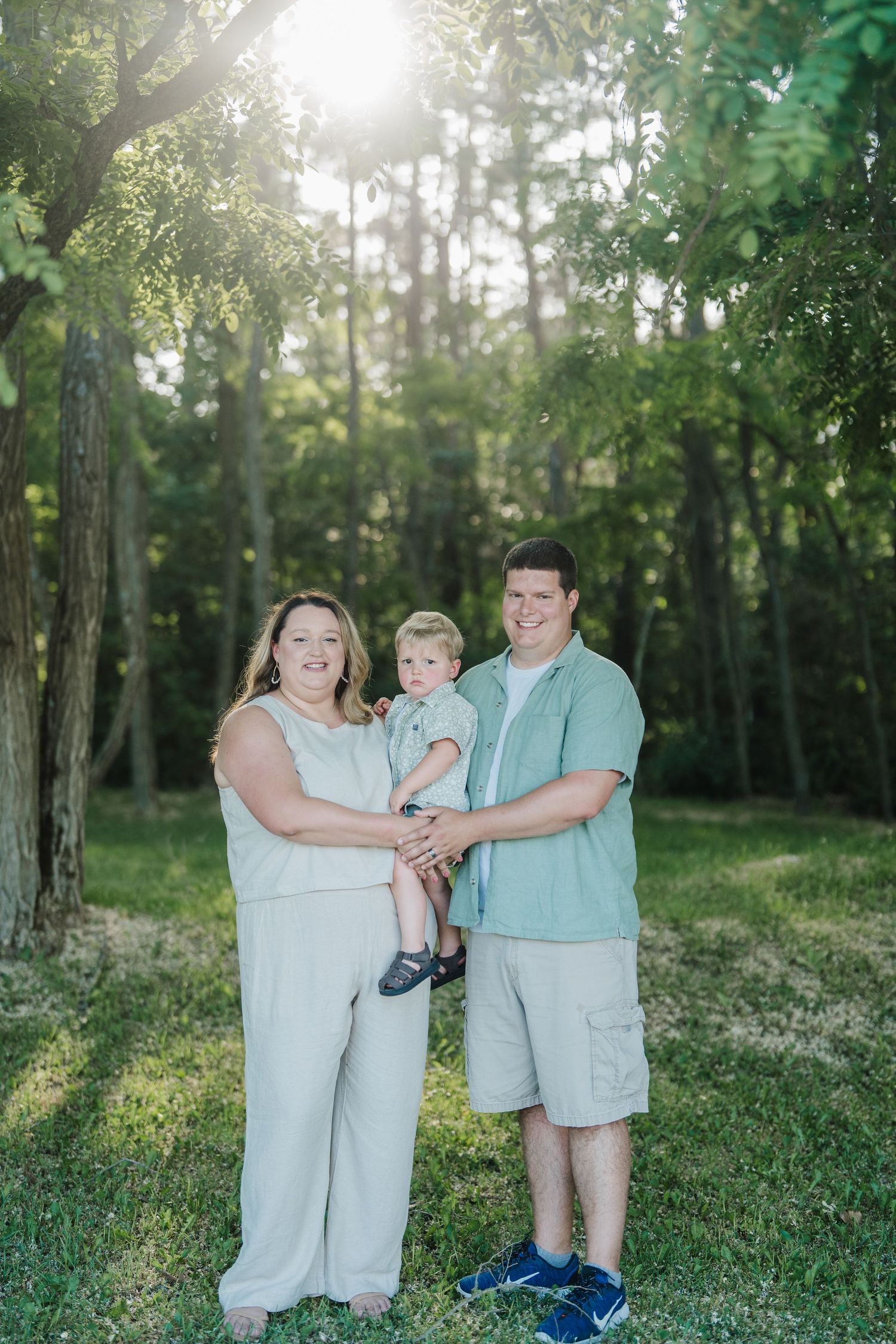 Family wearing coordinated mint green and white outfits poses together in a sunlit wooded area during golden hour.