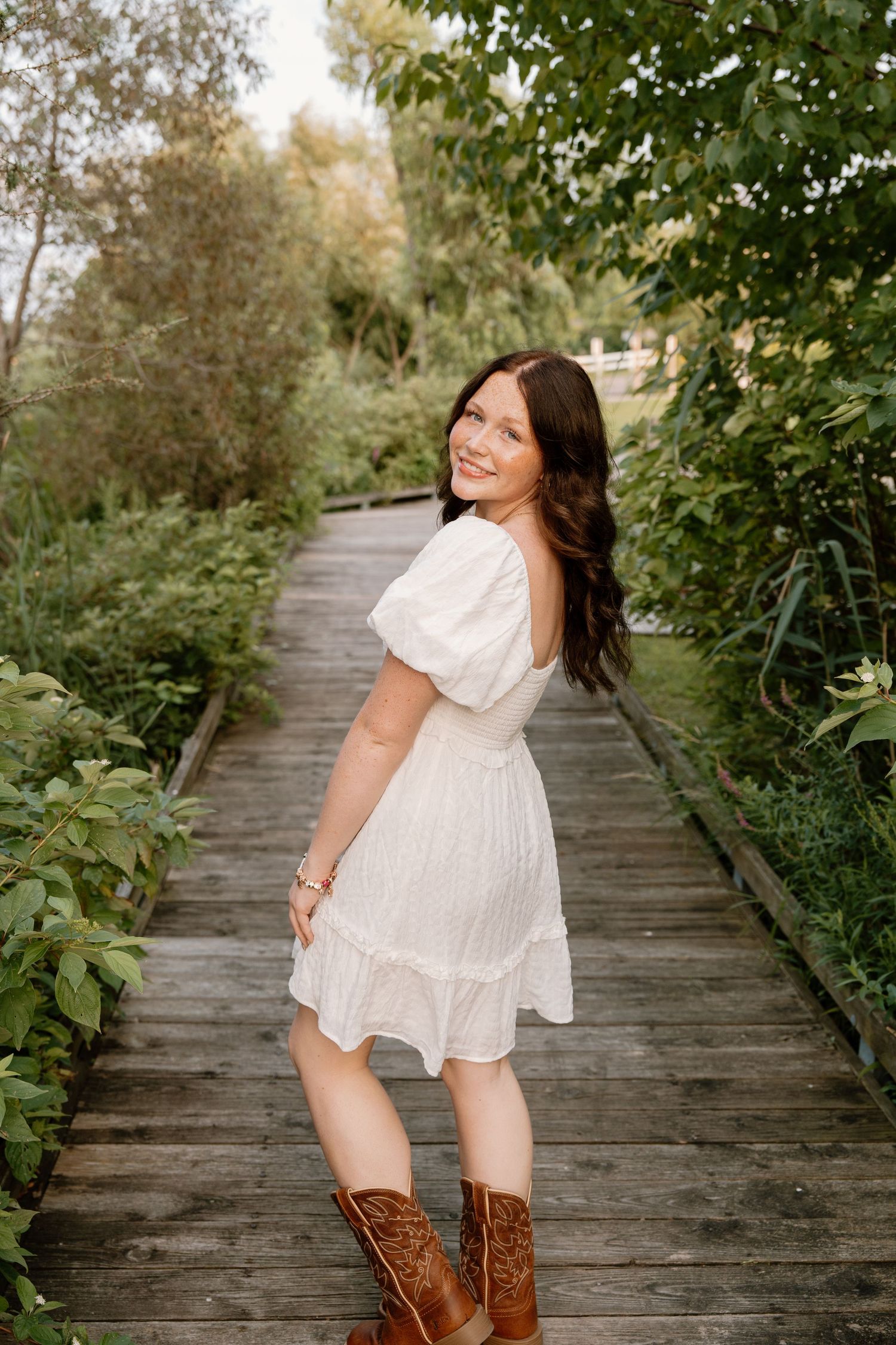 Young woman in white dress and cowboy boots smiling on a wooden boardwalk surrounded by lush greenery.