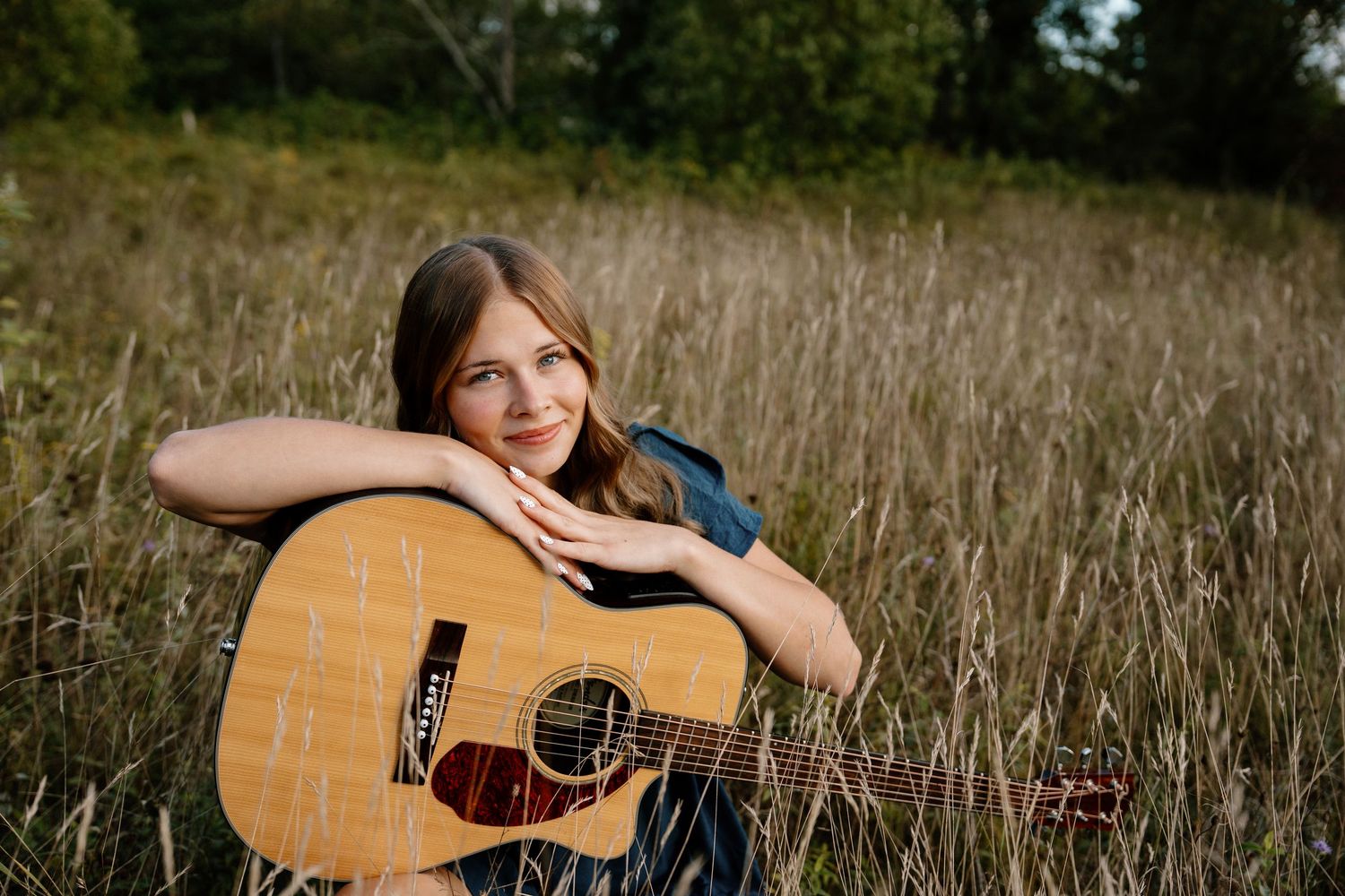 Young girl smiling in a field, leaning on an acoustic guitar surrounded by tall grass in golden hour light.