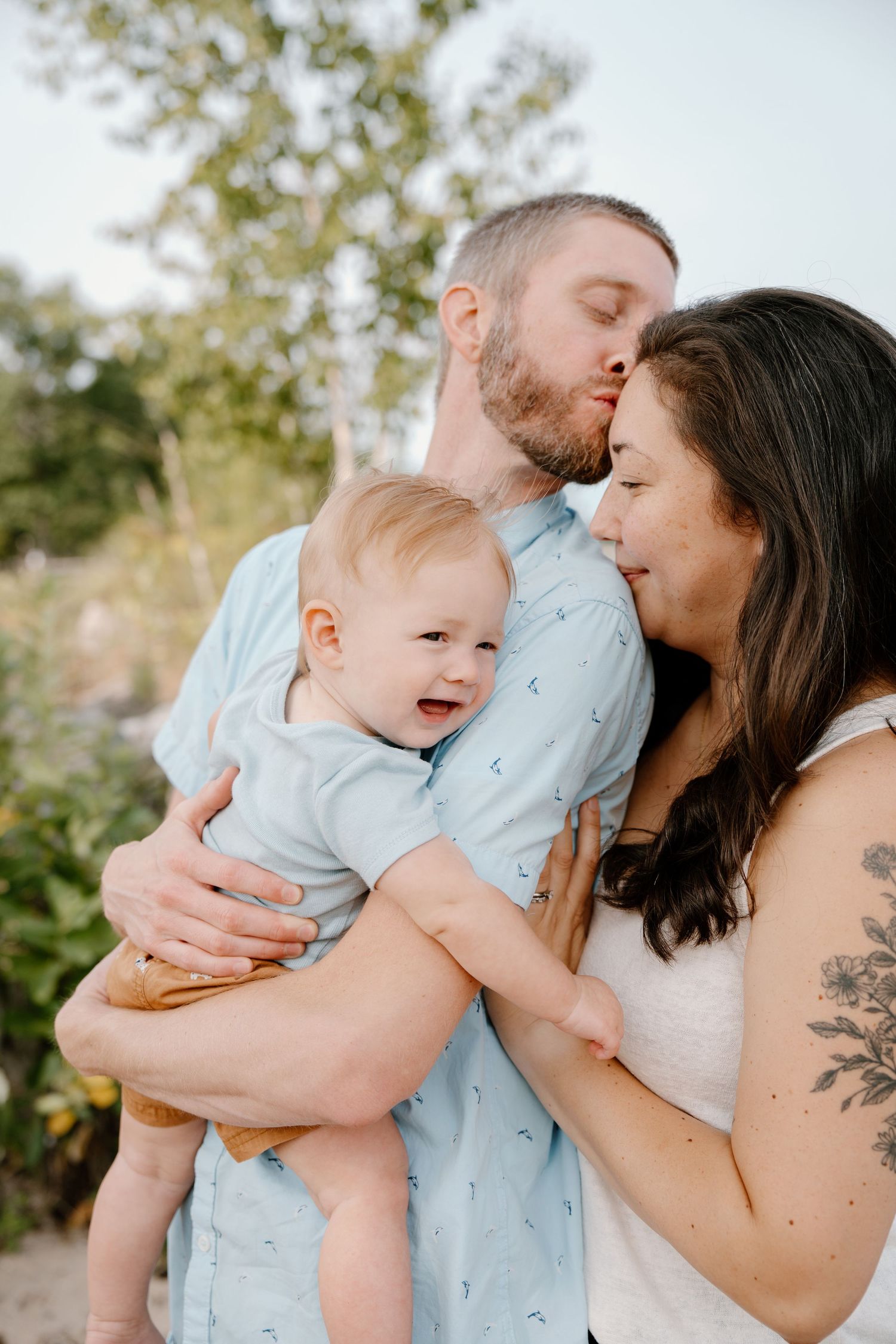 A couple shares a tender moment, dad kissing mom's cheek while holding their smiling baby outdoors.
