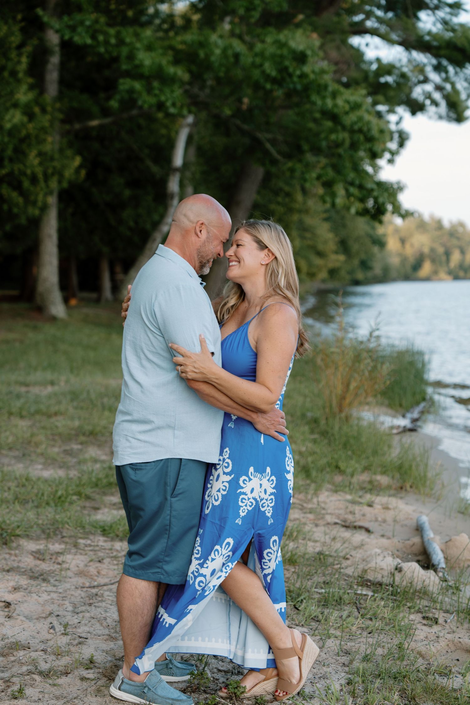 A couple embraces and smiles near a lakeside beach, woman in blue floral dress, man in light shirt and shorts.