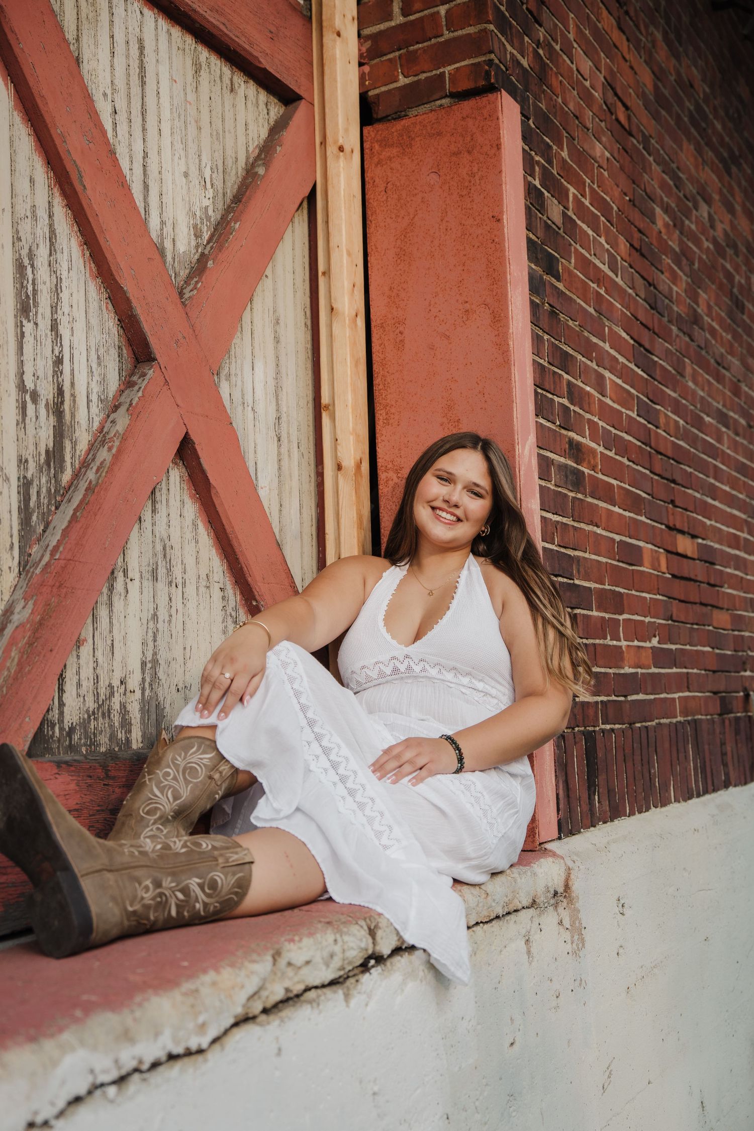 Young woman in white lace dress and cowboy boots sitting by rustic brick wall and barn door, smiling.