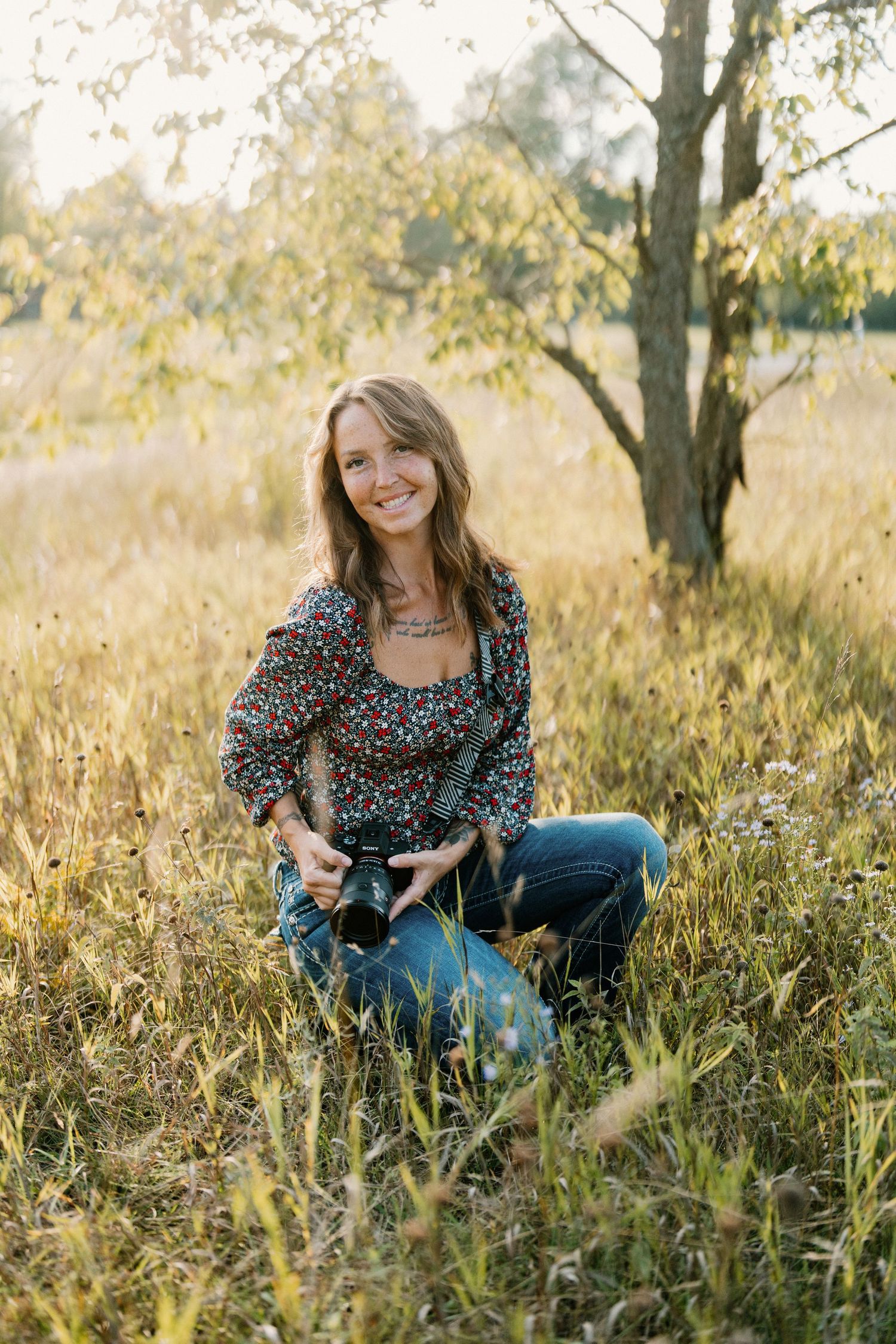 Smiling woman holding a camera sits in a sunlit grassy field wearing a floral top and jeans near a tree.