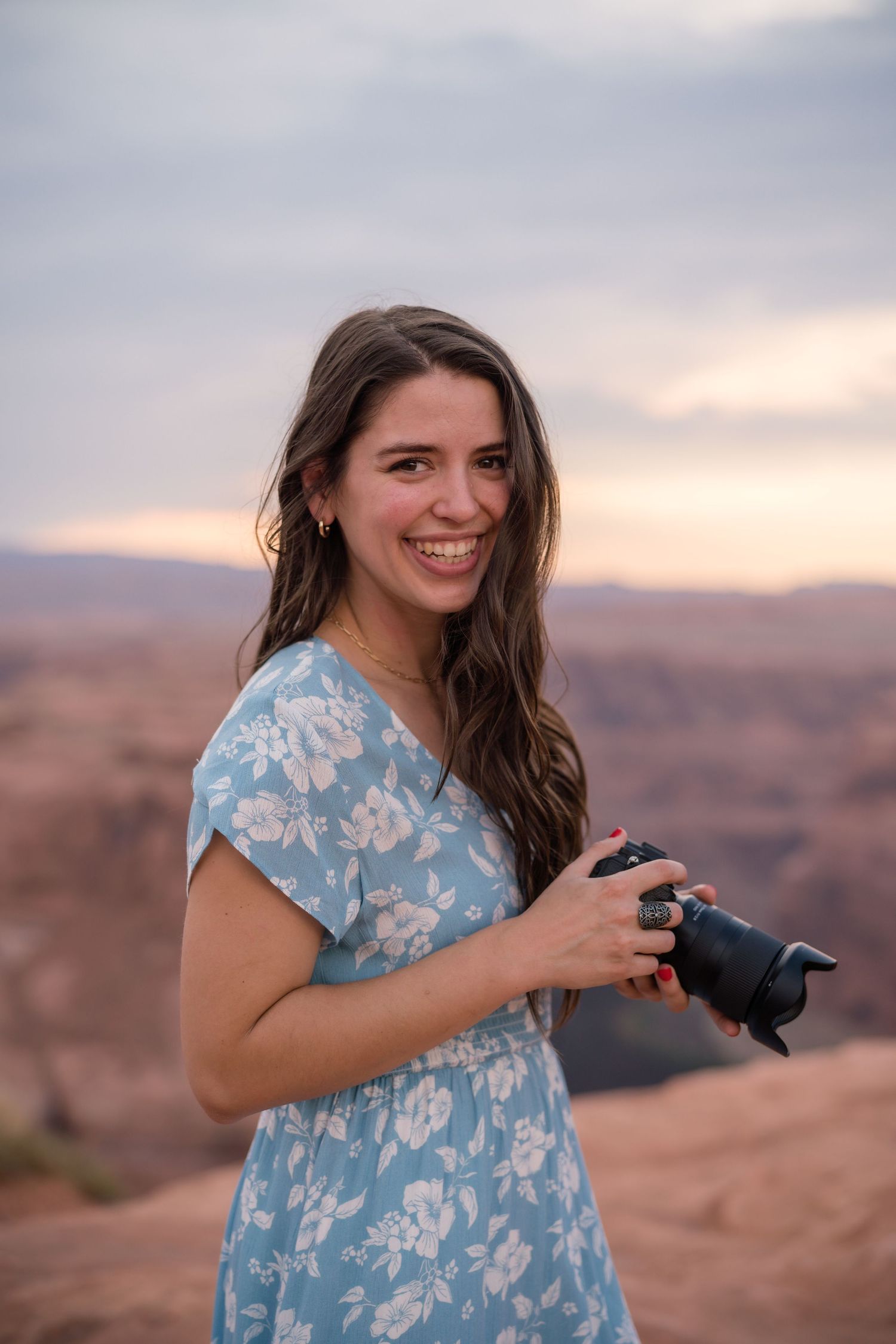 Smiling woman in floral blue dress holding a camera at a scenic canyon overlook during sunset.