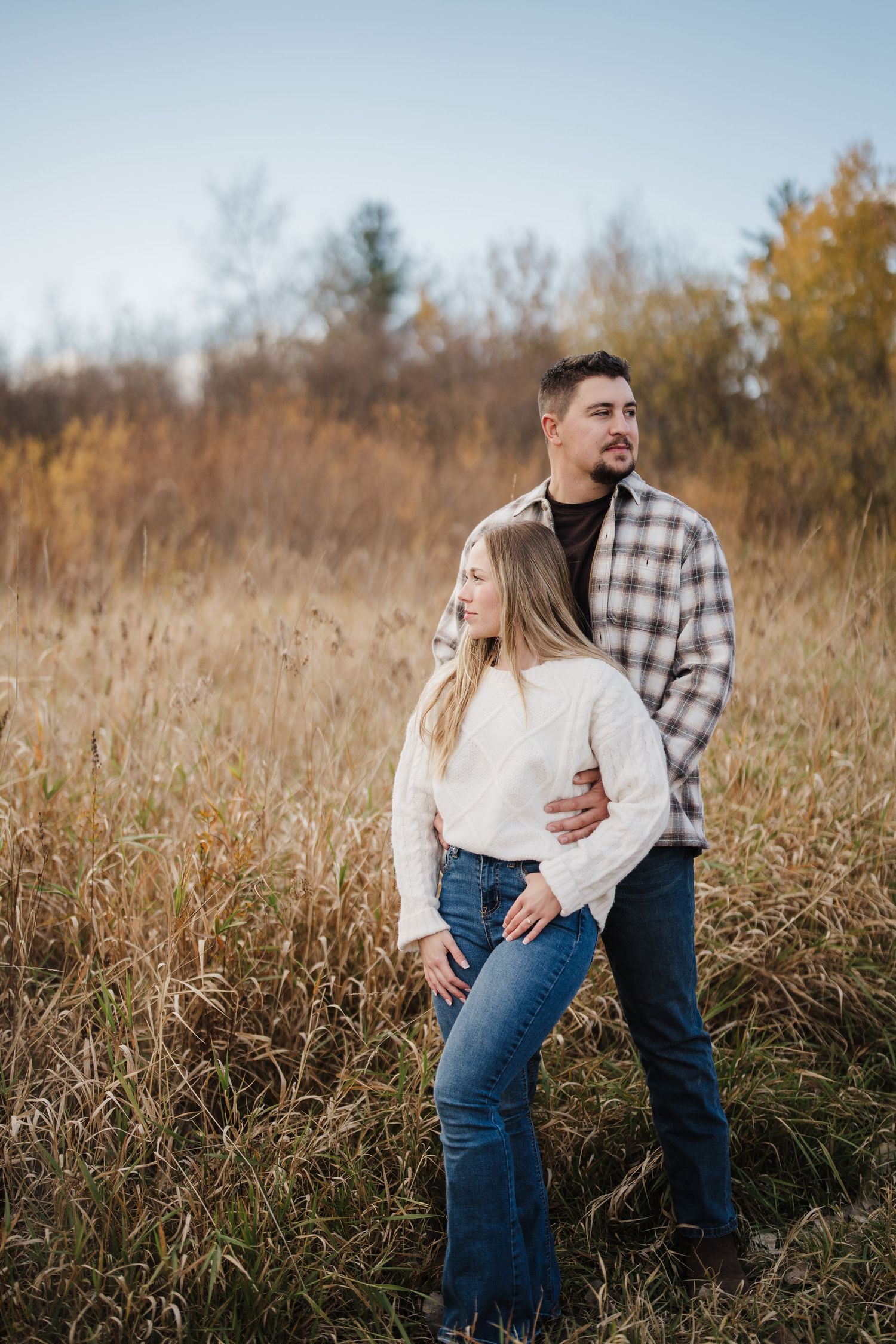 Couple embraces in a golden autumn field with tall grass and trees in the background during sunset.