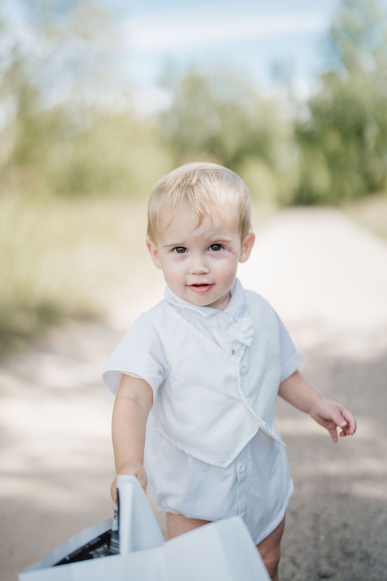 Young child in white clothing sits on a sandy surface with bright sunlight and trees in the background.