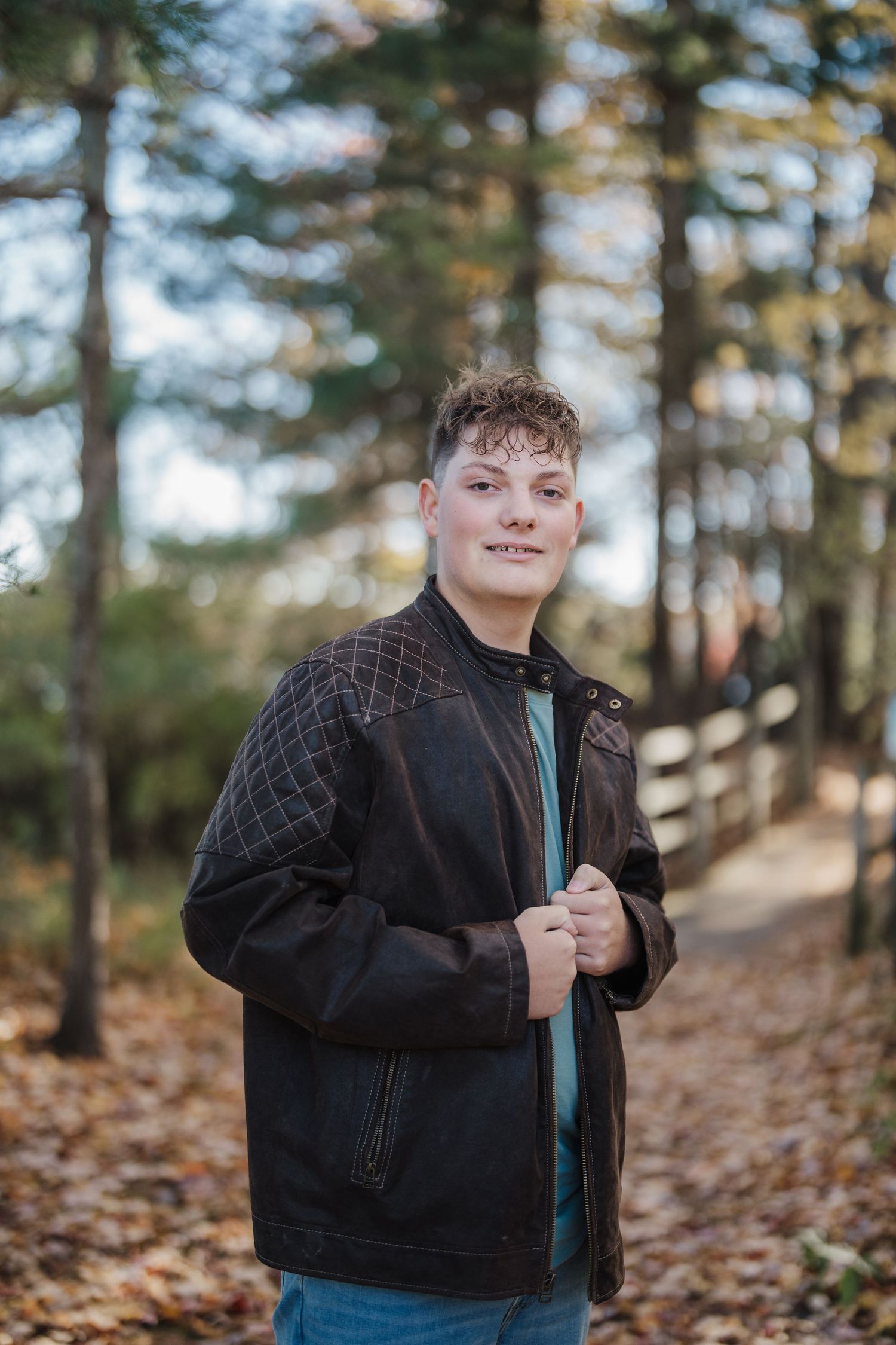 A person wearing a black denim jacket poses against a wooded autumn background with fallen leaves and pine trees.