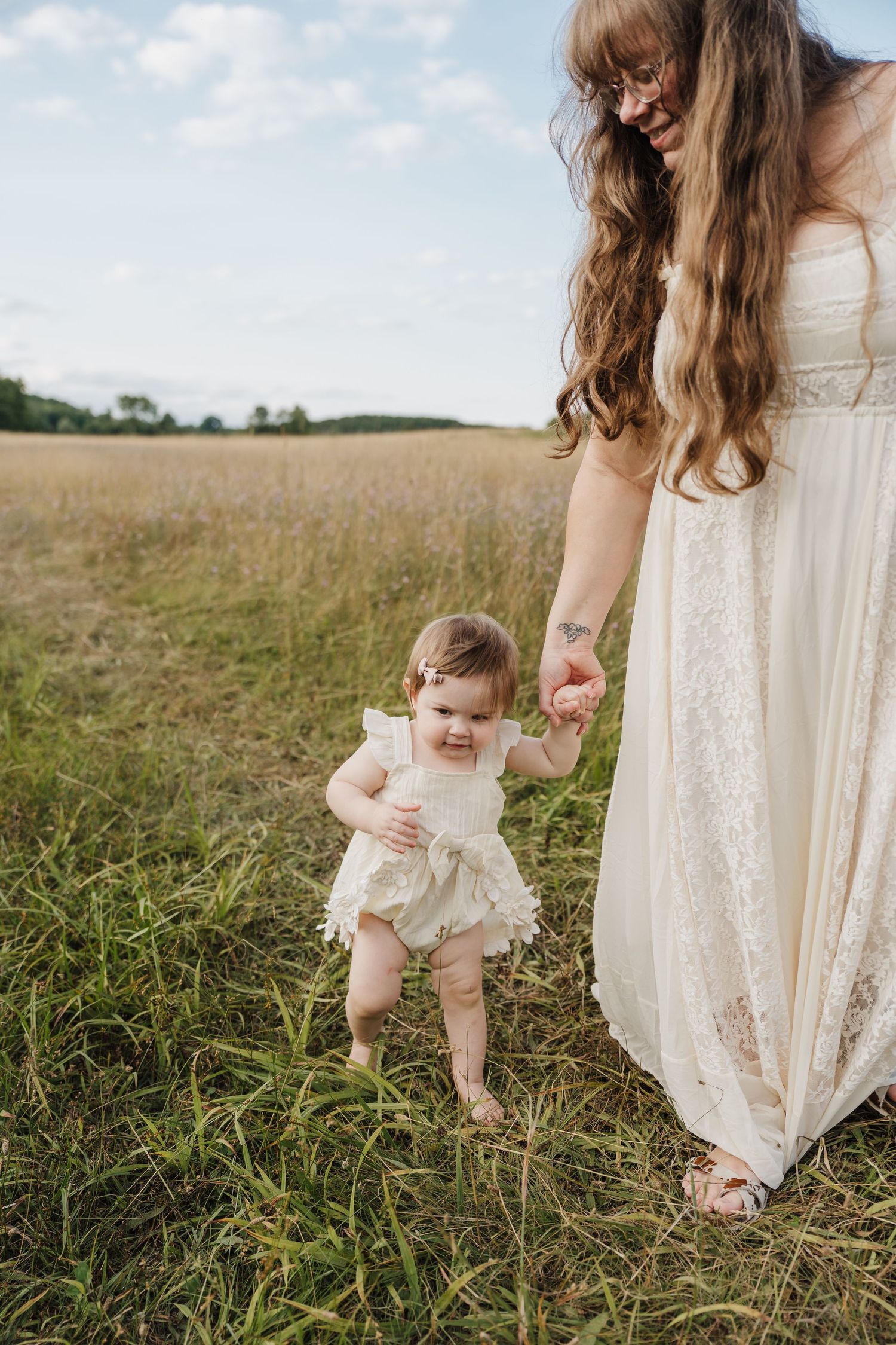 A mother in a white lace dress helps her baby girl take first steps in a sunny green field.