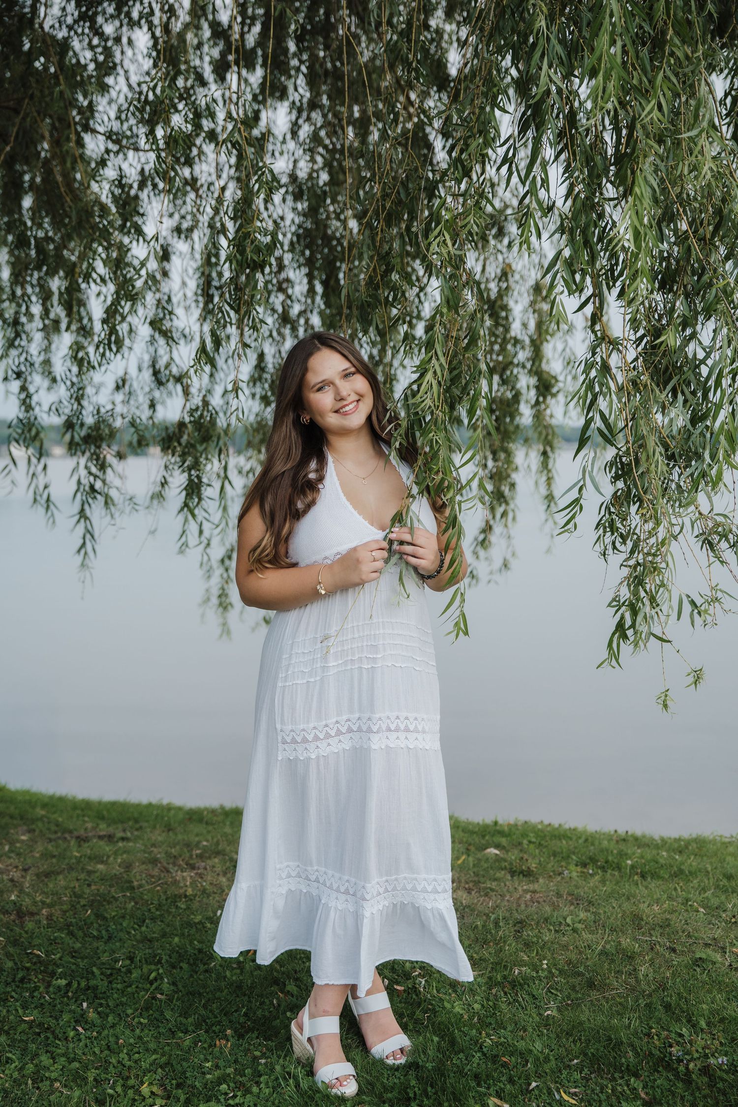 Young woman in white sundress standing under weeping willow tree by lakeside, smiling outdoors.