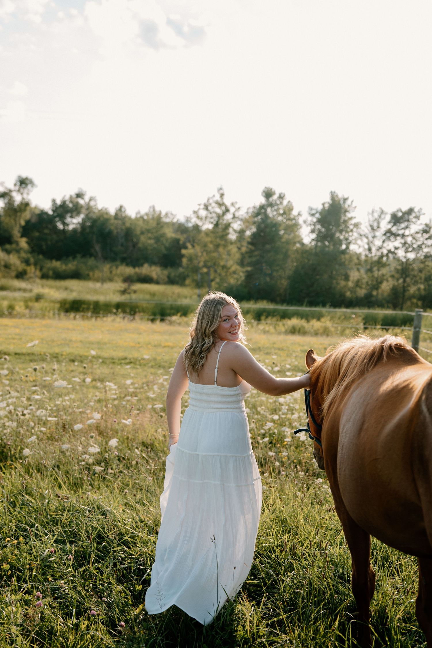 Woman in white maxi dress smiling while petting a chestnut horse in a sunny green field.