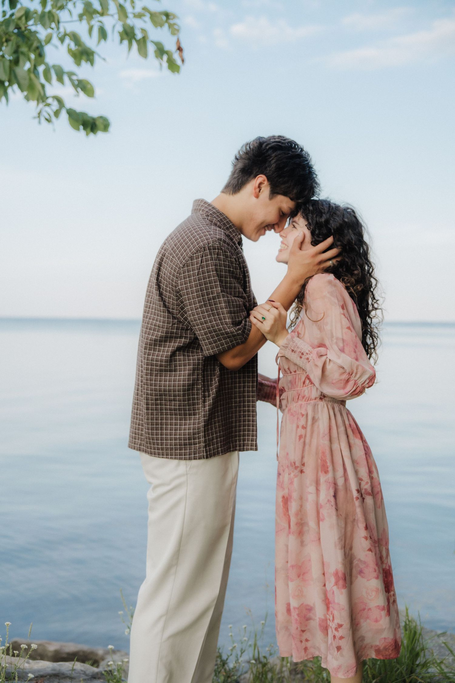 A couple shares a romantic moment by the water, forehead to forehead, dressed in floral and plaid attire.