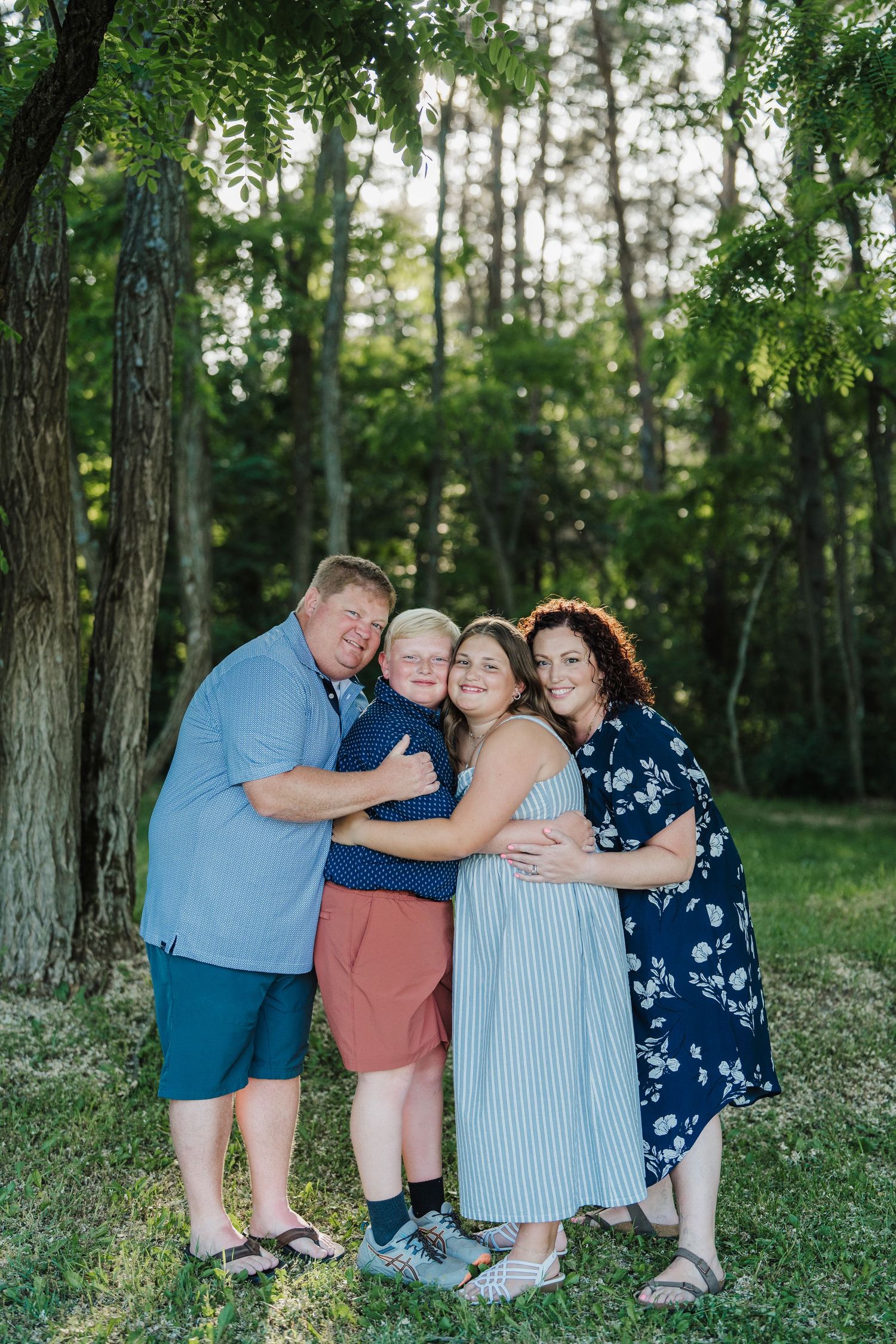 A family group in coordinating blue and pink outfits poses playfully together on grass in a wooded outdoor setting.