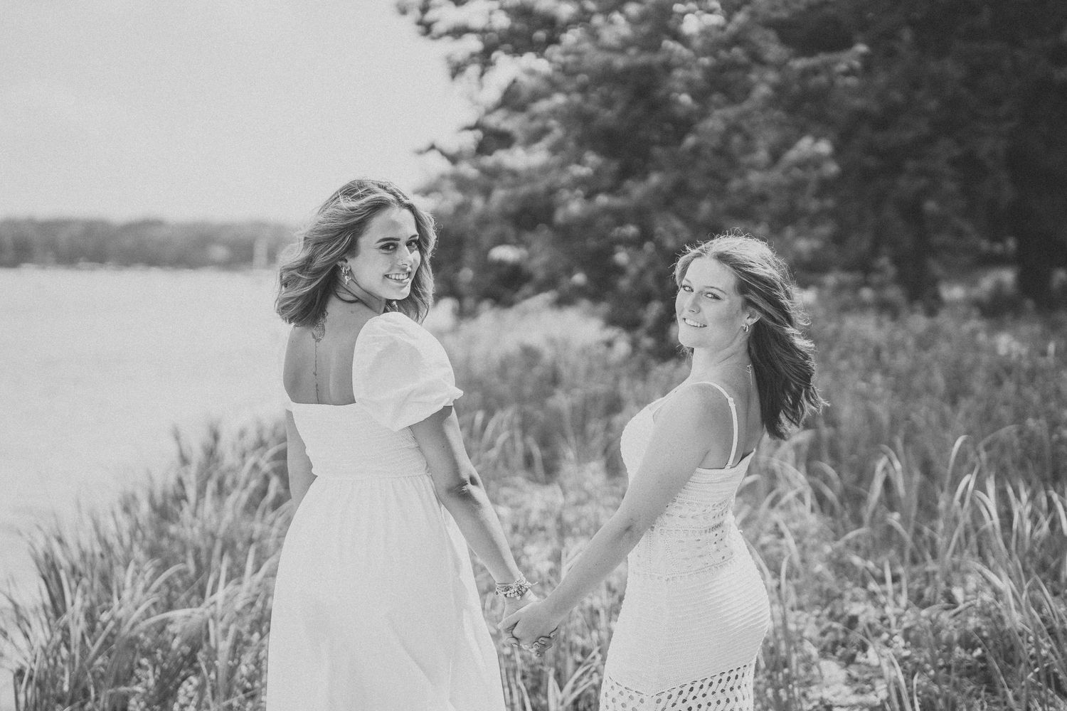 Two women in white dresses pose together by a lakeside in a black and white photograph.