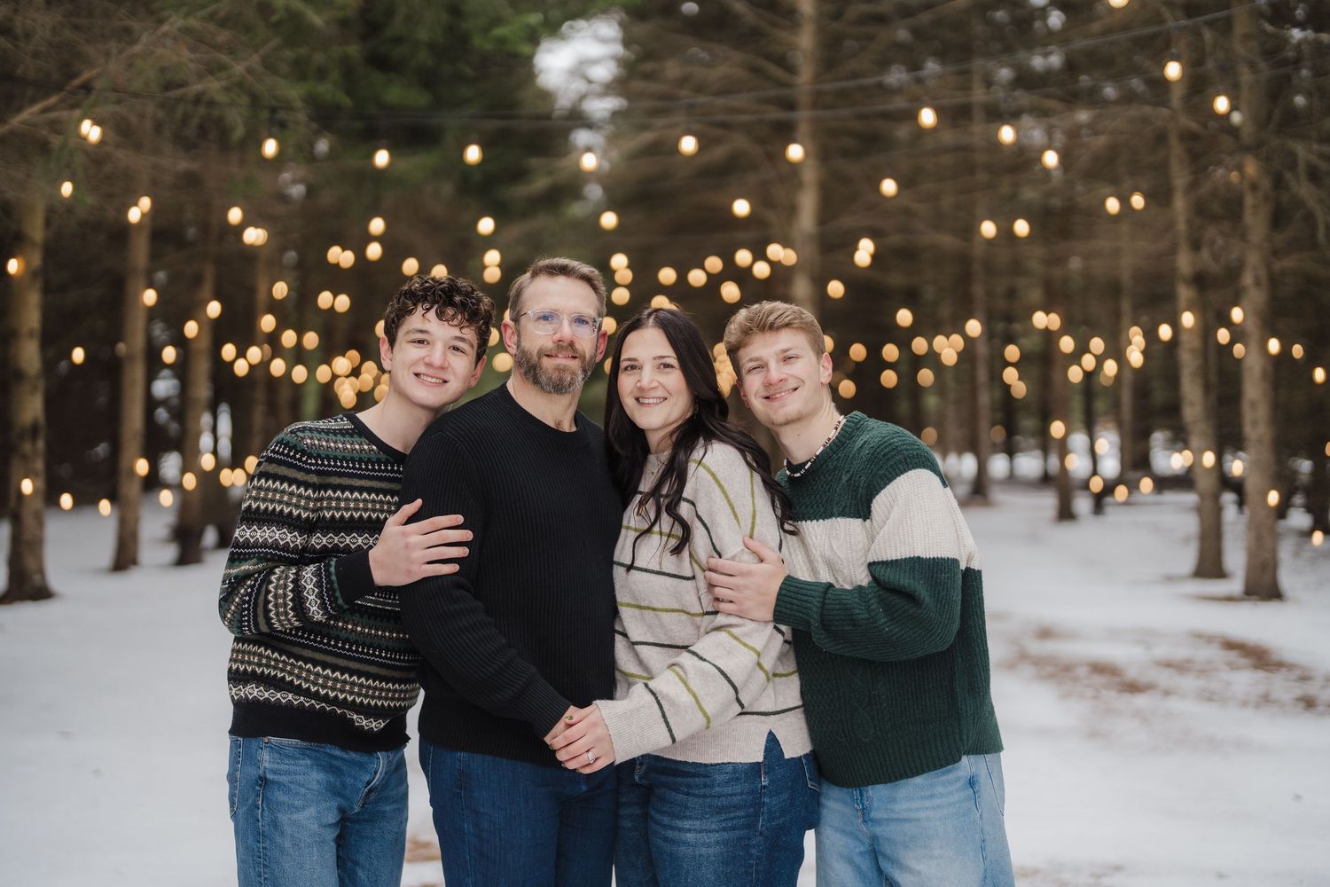 Family members embrace while seated on a rustic bench surrounded by snow-covered trees and glowing string lights.