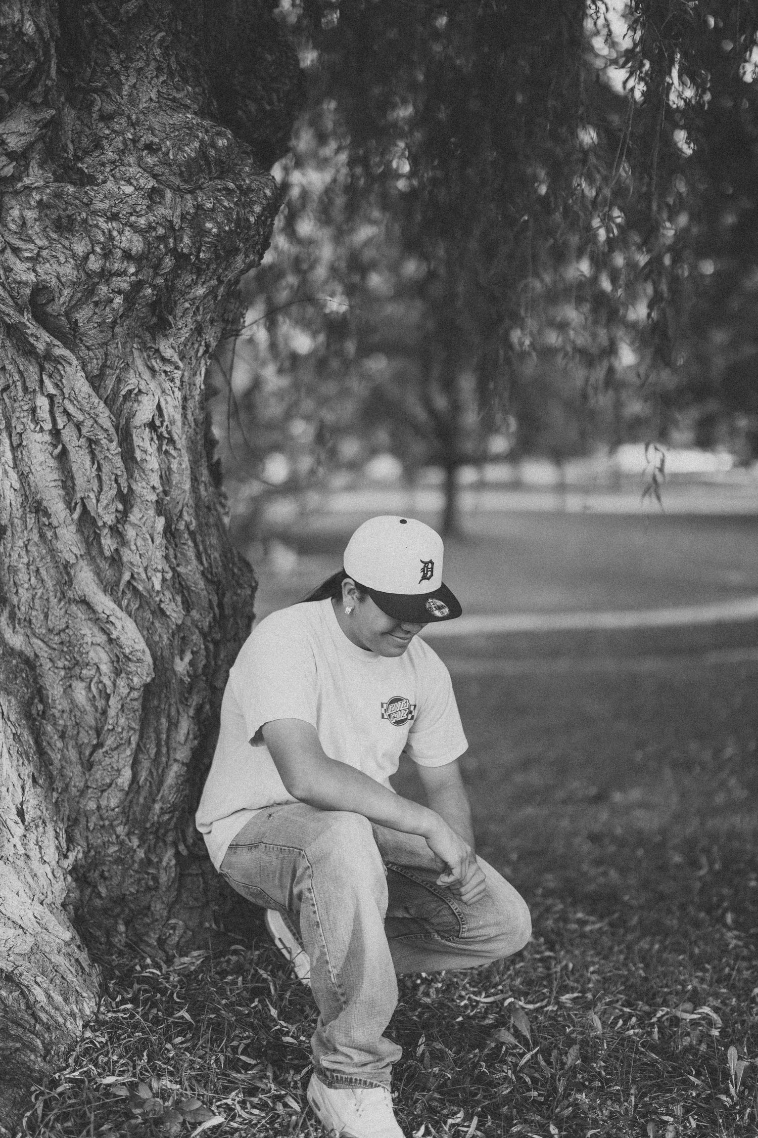 Black and white portrait of someone in a baseball cap sitting by a large tree trunk in a park.