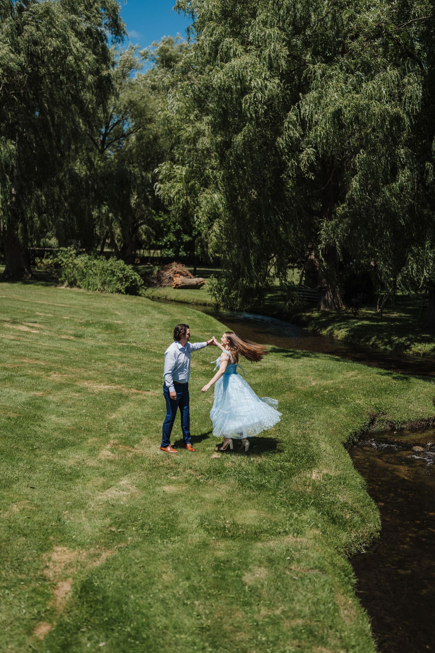 A couple dances together on a grassy slope surrounded by willow trees in a romantic outdoor setting.