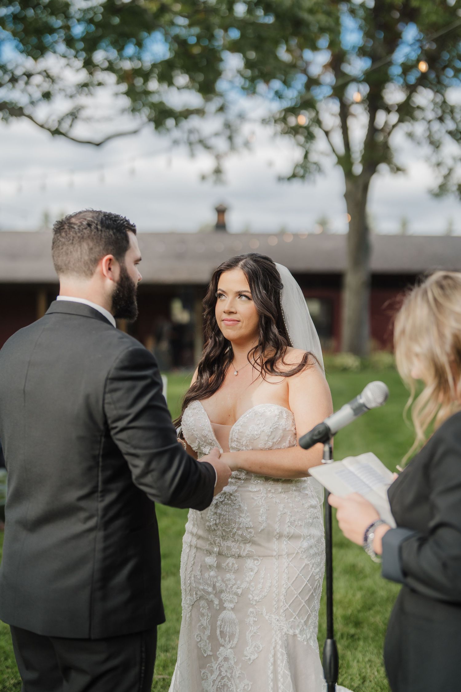 A wedding couple stands outdoors in a grassy area with trees while others gather around them.