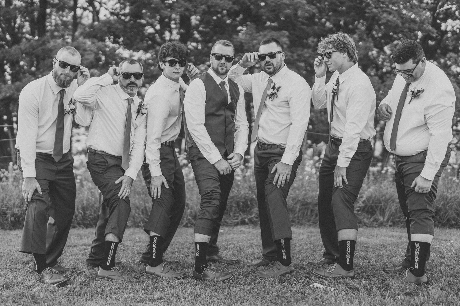 A line of groomsmen in matching gray suits pose together outdoors at a wedding ceremony.