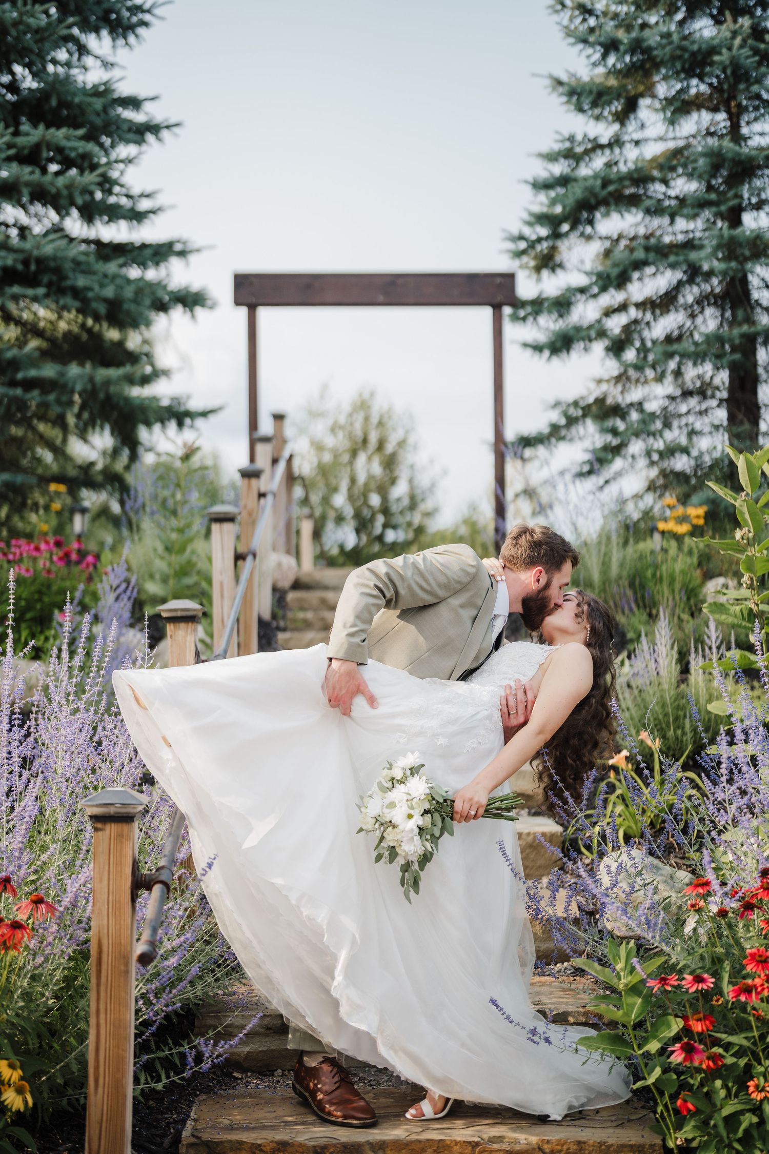 A romantic dip kiss between newlyweds on a wooden bridge surrounded by colorful garden flowers.