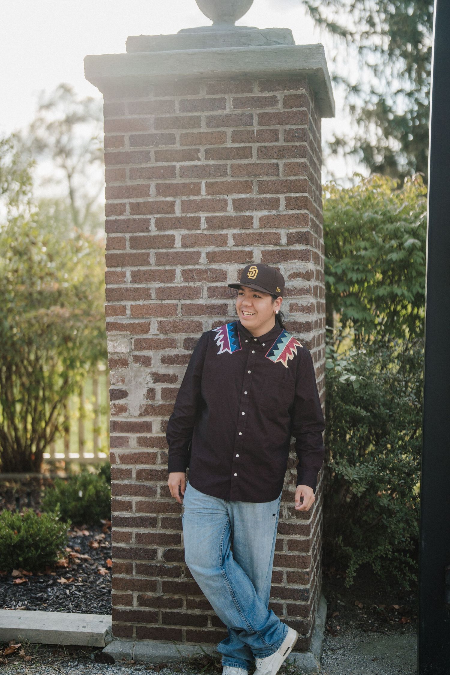 A person wearing a black button-up shirt and jeans poses casually against a brick wall in an outdoor setting.