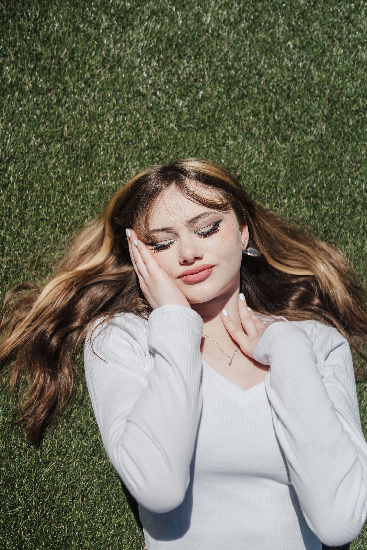 Person in white sweater lies on green grass during an outdoor photo session with natural lighting.