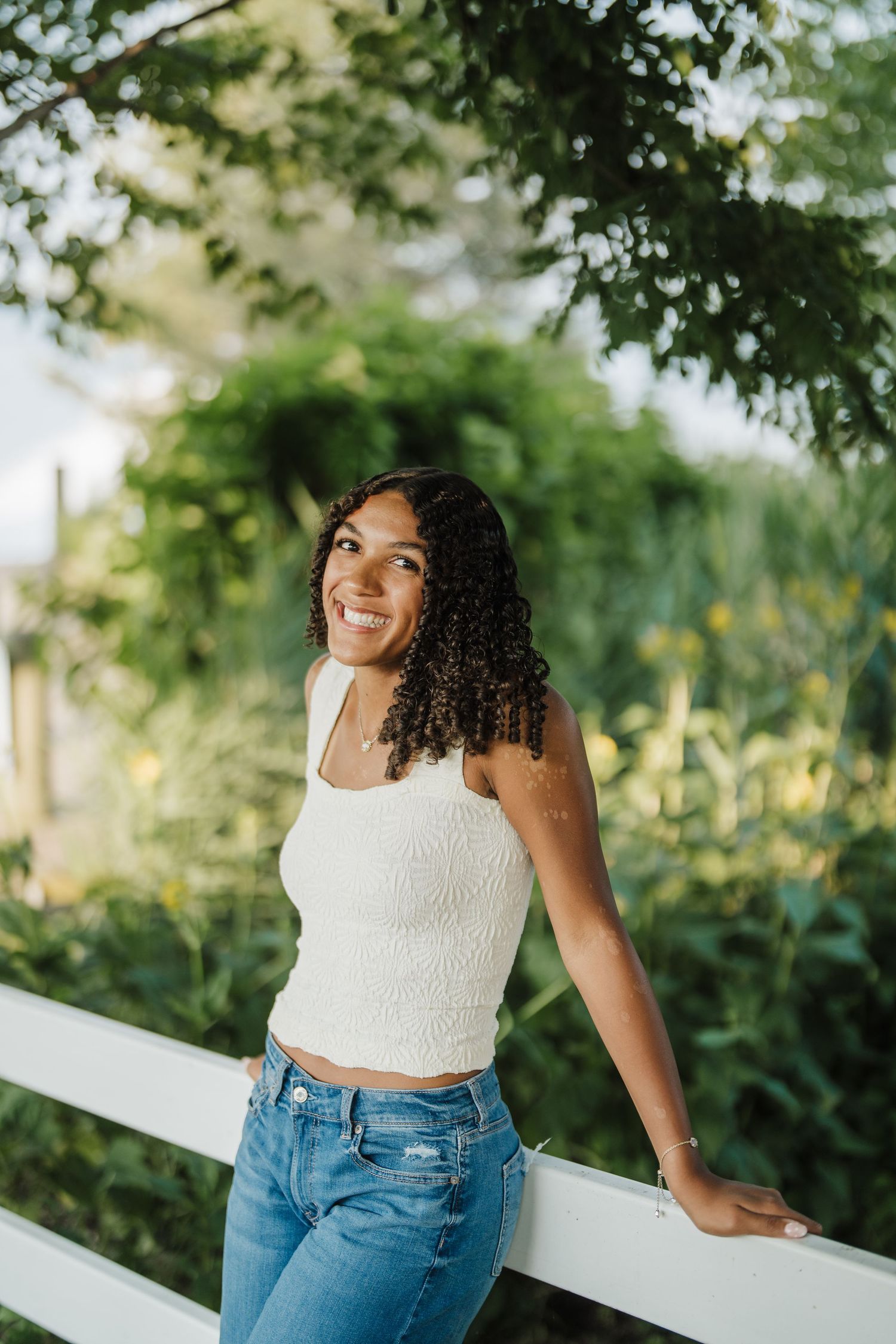 A person in a white crop top and jeans leans against a white railing outdoors with lush greenery in the background.