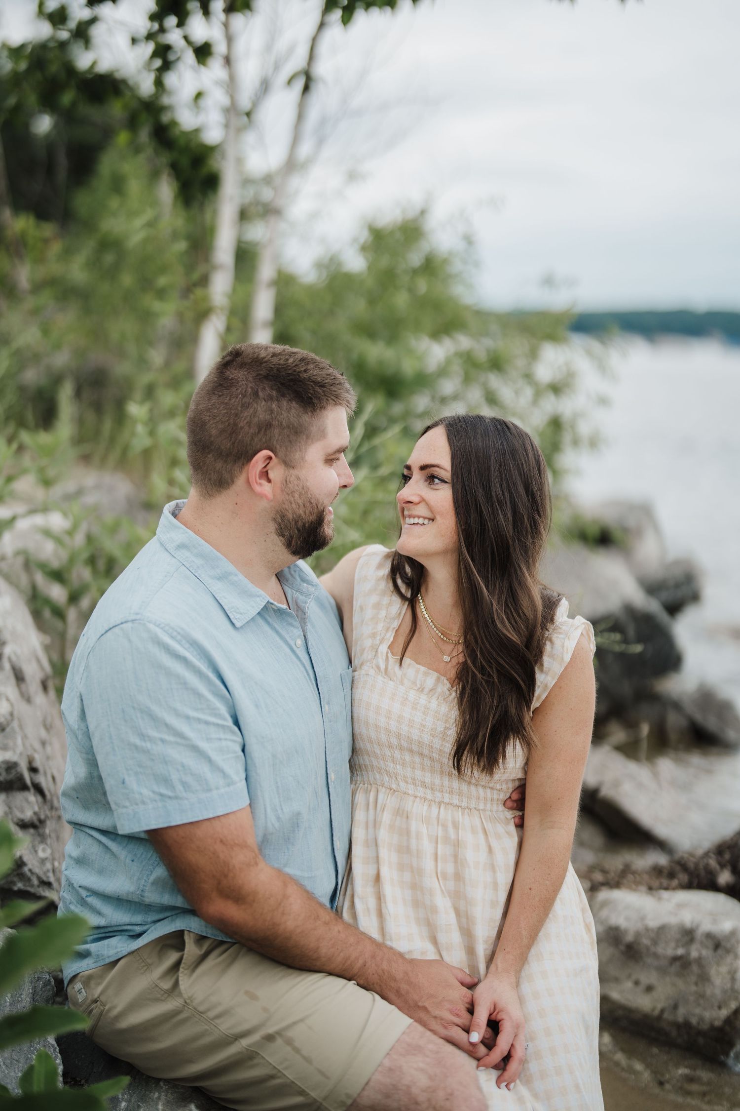 A couple shares tender moments together while sitting on rocky shores near a lake on a bright summer day.