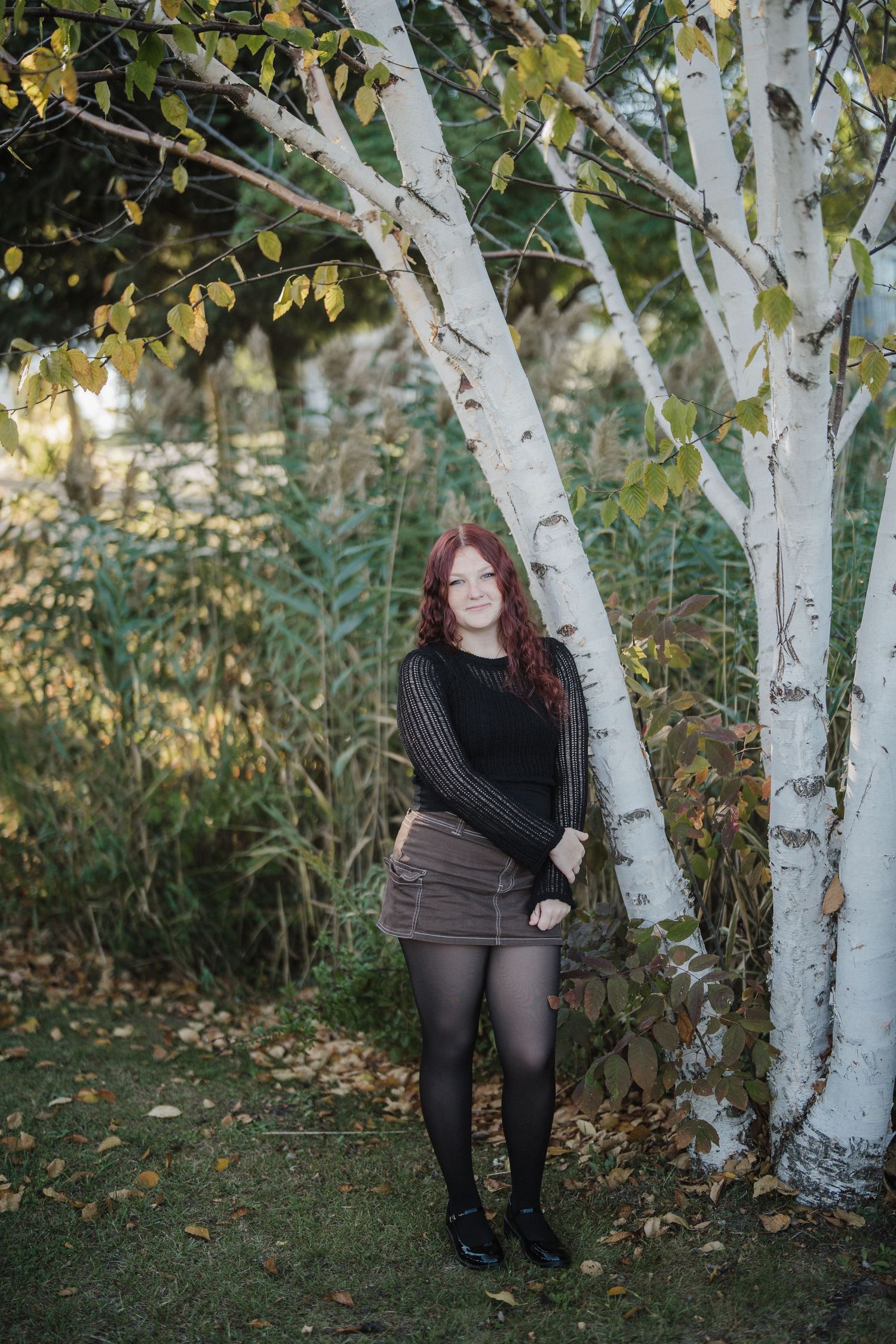 A figure in a black sweater and brown skirt stands beside white birch trees in an autumn garden setting.