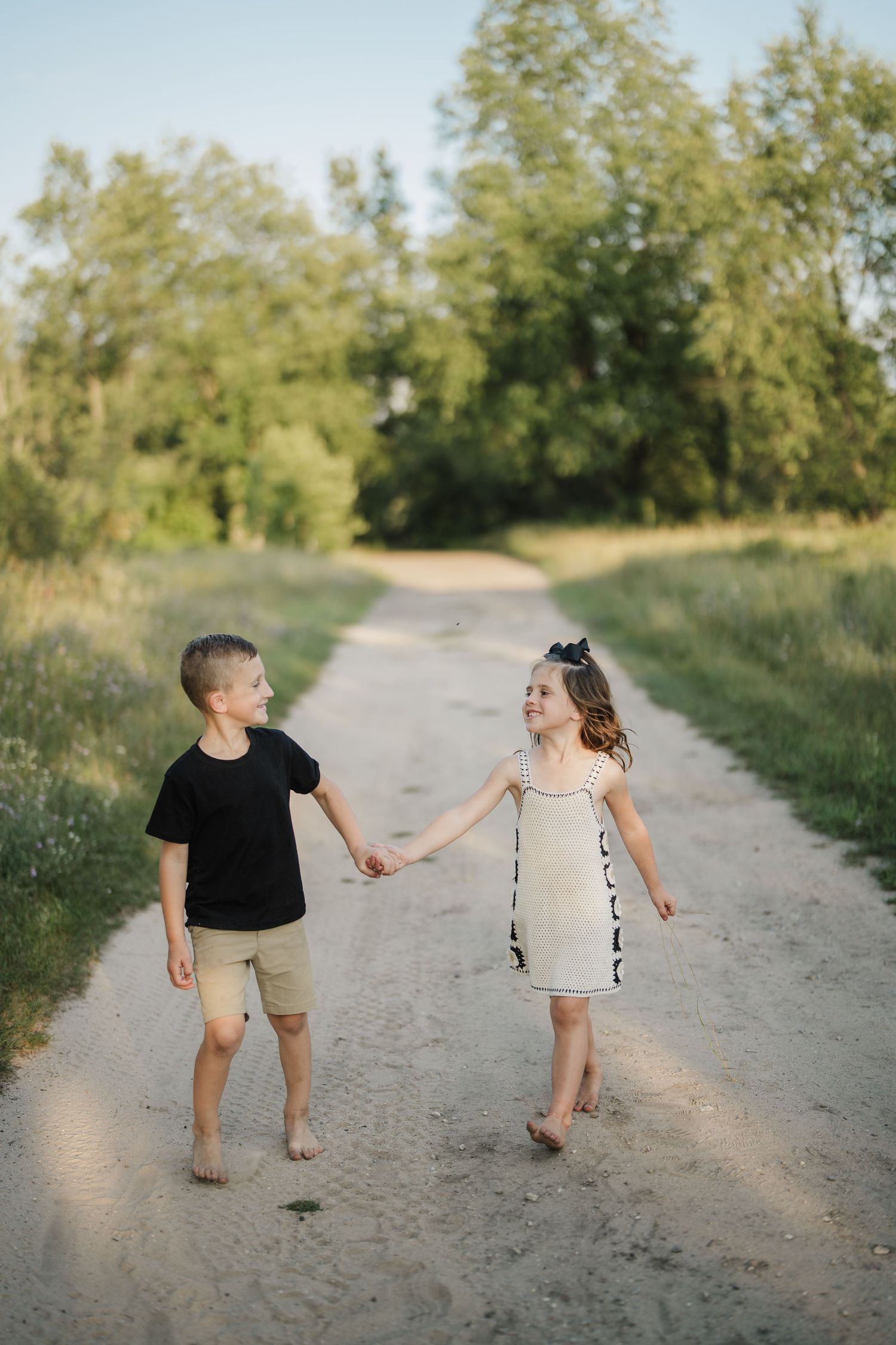 Two children hold hands while walking barefoot down a dirt path surrounded by green trees.
