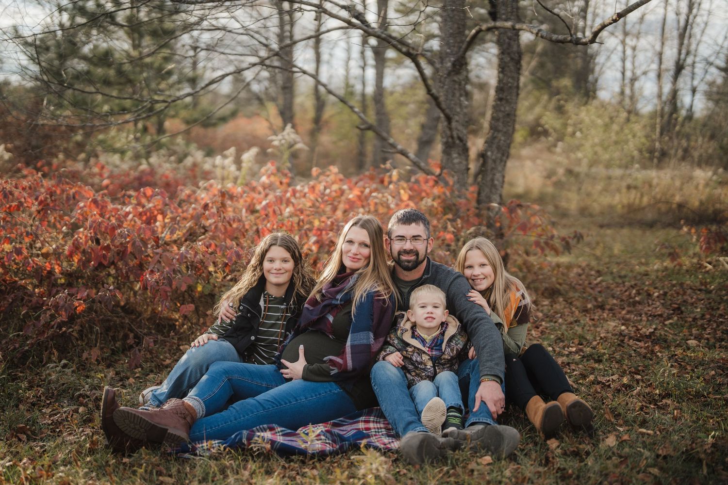 A group sits together on fallen autumn leaves in a wooded area during a casual outdoor photo session.