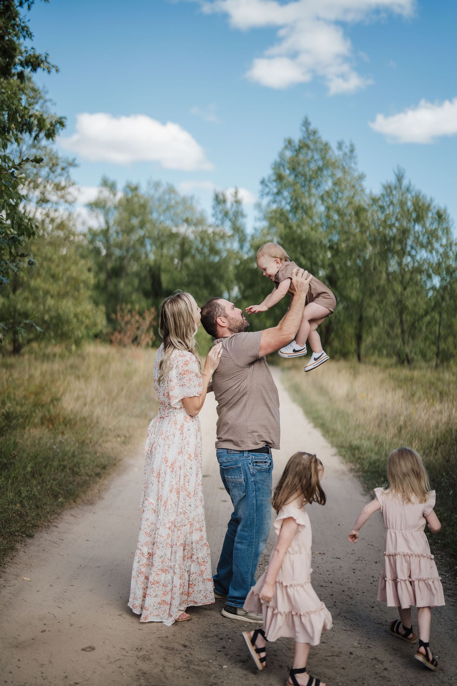 A family walks together on a dirt path surrounded by green trees on a sunny day with white puffy clouds in the sky.