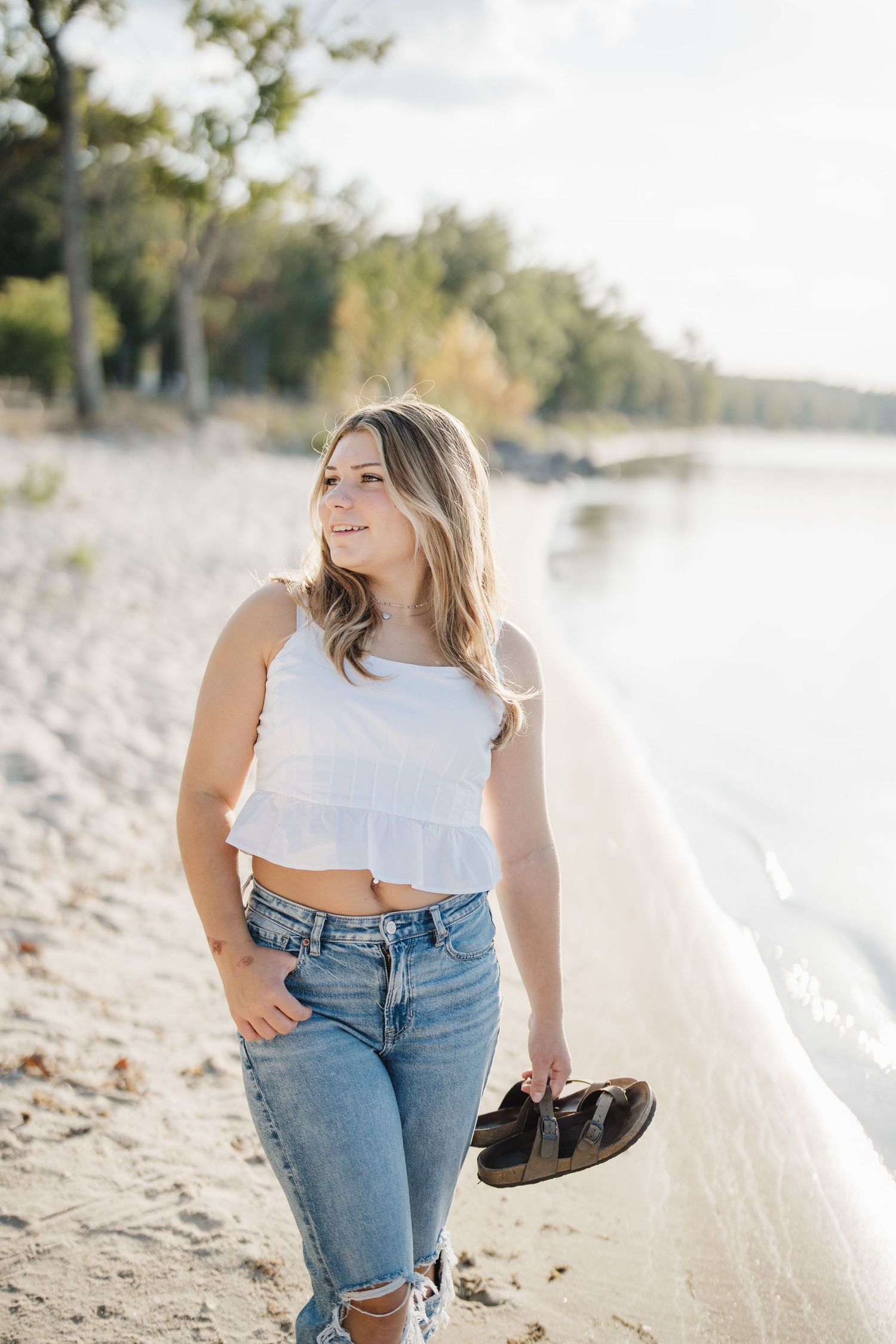 A person in a white crop top and jeans poses casually on a sandy beach during golden hour.