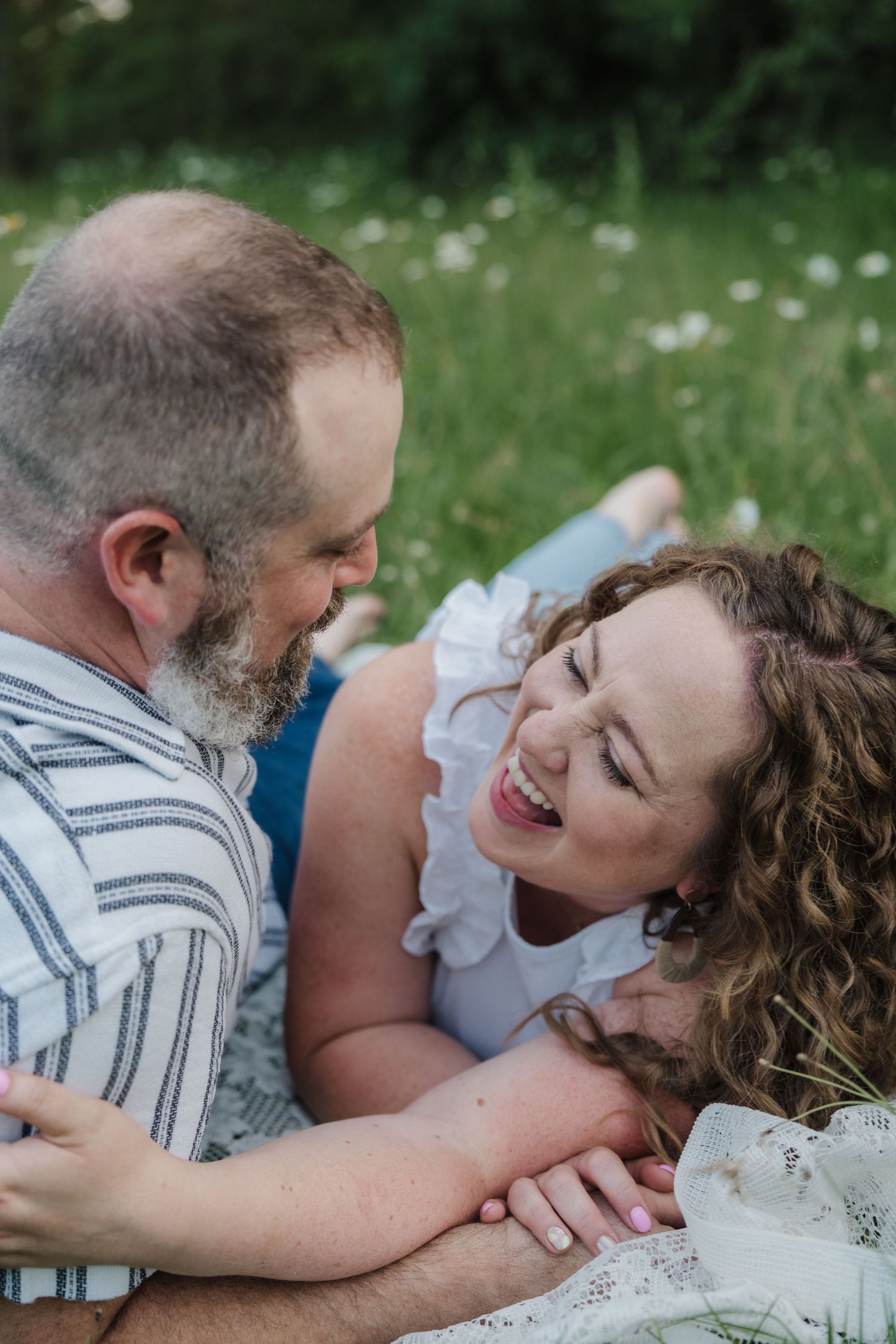 Happy couple laughing and gazing at each other while lying on a blanket in a daisy-filled green meadow.